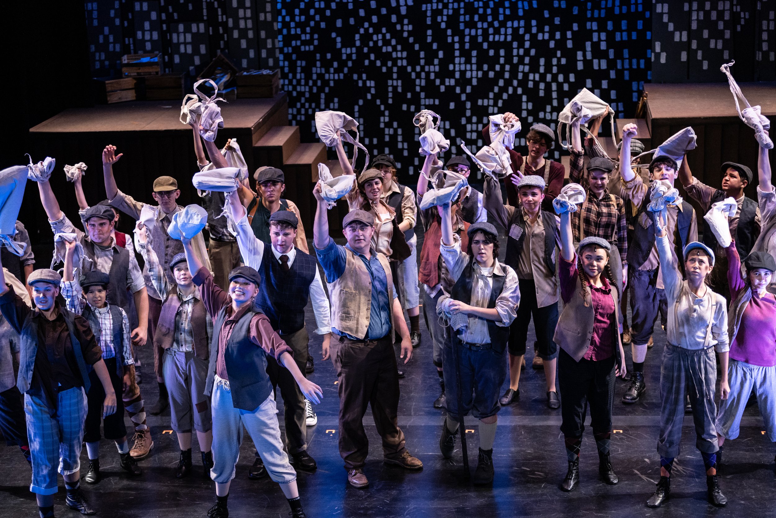A group of young performers dressed in vintage-style costumes, including caps and vests, performing on stage with a backdrop displaying digital or pixelated patterns. Some are holding cloth bags and appear to be part of a theatrical or dance producti