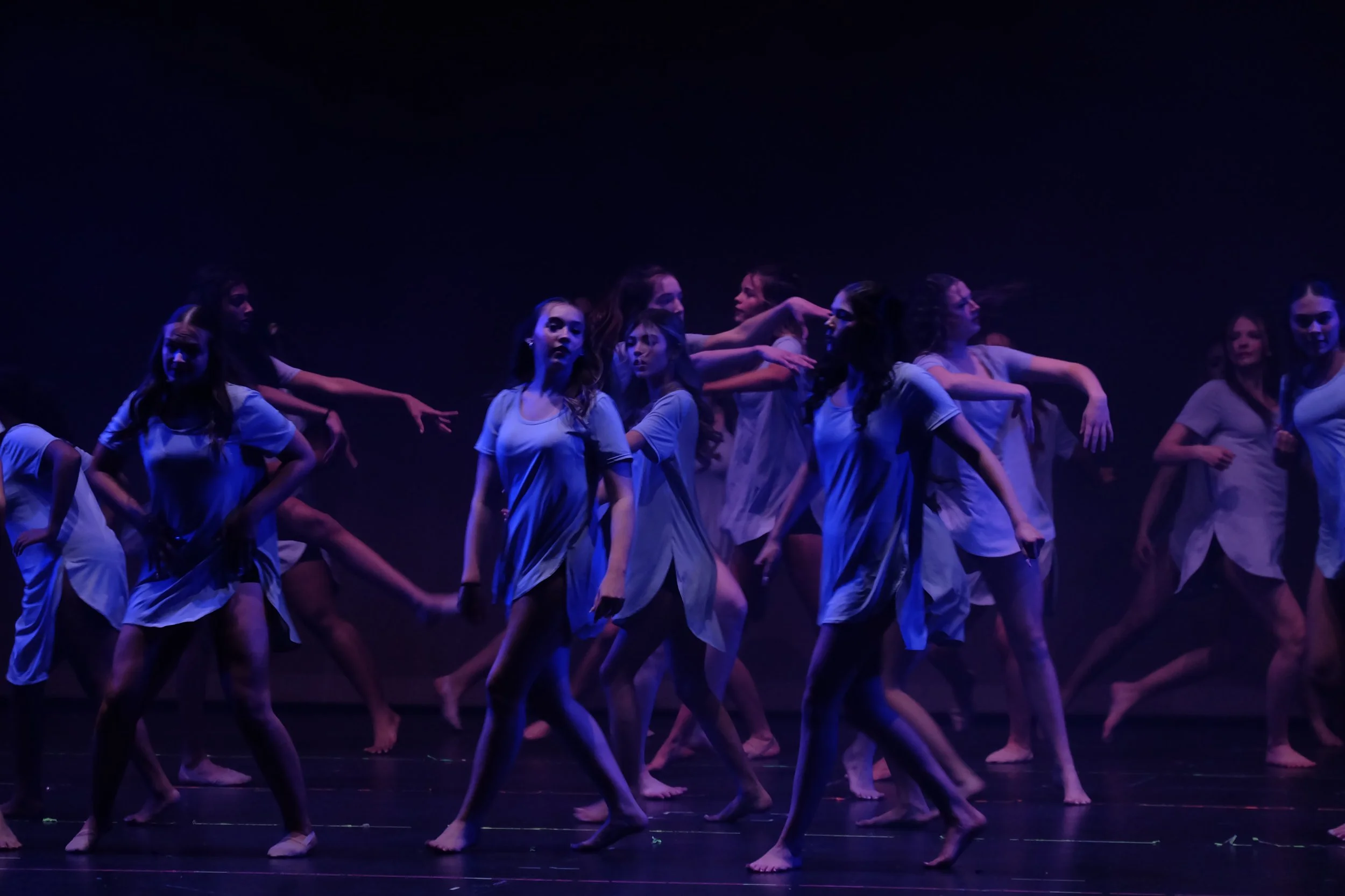 Group of dancers performing on stage under dim purple lighting, wearing light-colored clothing.
