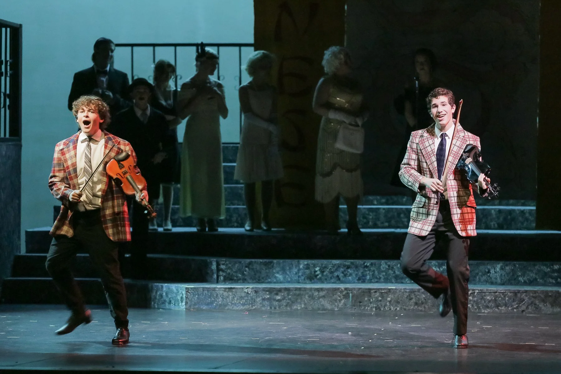 Two male performers in plaid blazers and dress shoes dancing on stage, with a group of behind-the-scenes actors in elegant costumes observing from the stairs in the background.