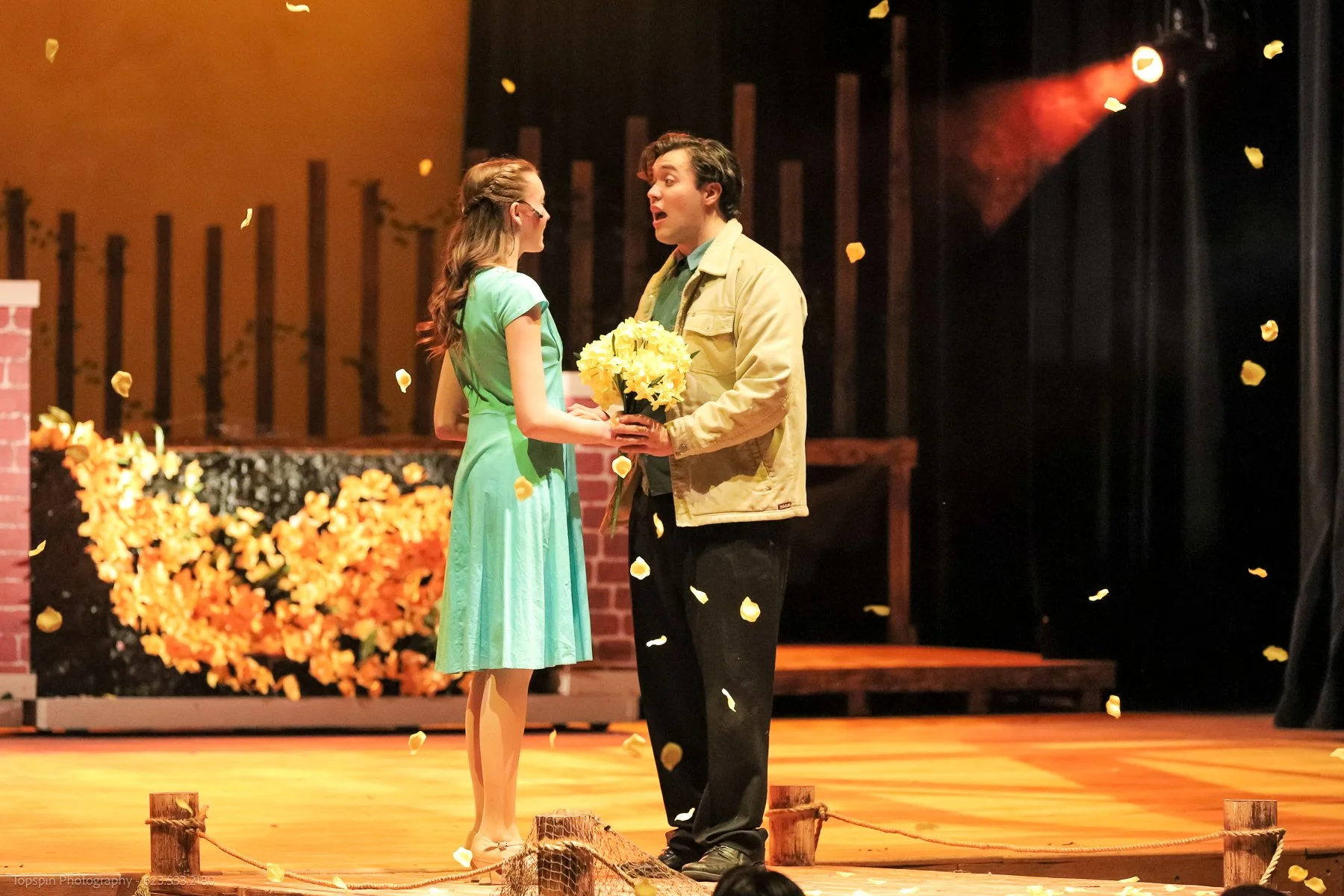 A young man and woman on stage during a theatrical performance. The woman, wearing a green dress, and the man, in a beige jacket, are sharing a moment while the man presents her with a bouquet of yellow flowers. Yellow petals are falling around them, and the stage background includes orange flowers and a brick wall.