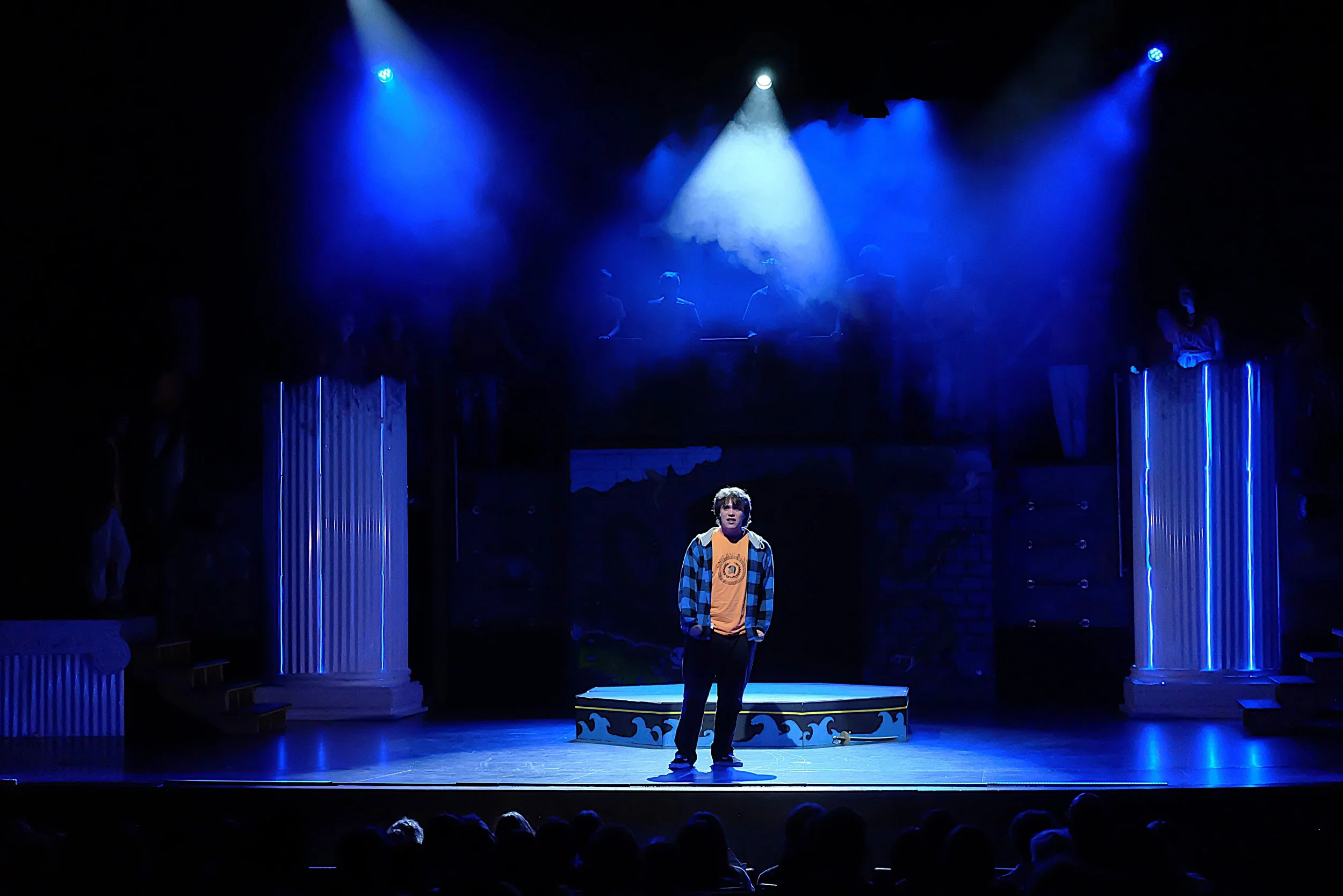 A person standing on a stage illuminated by blue and white stage lights, with a set design featuring columns and a platform. Several other people are visible in the background, some on the platform and others off to the sides, with an audience watchi