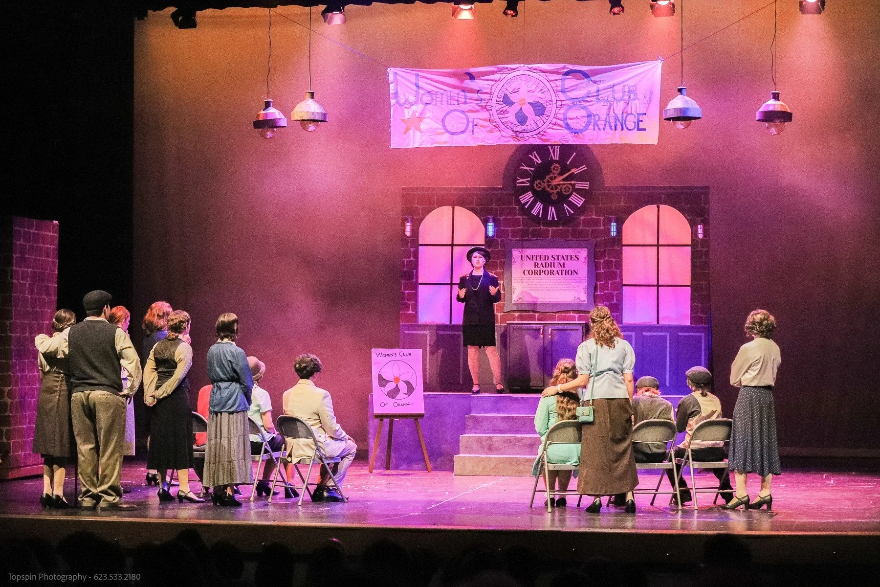 The image depicts a theatrical stage with a group of actors. A woman standing on a platform appears to be speaking to the audience. The background shows a clock, a brick wall, and banners, including one that reads 'Women's Club of Orange' and another
