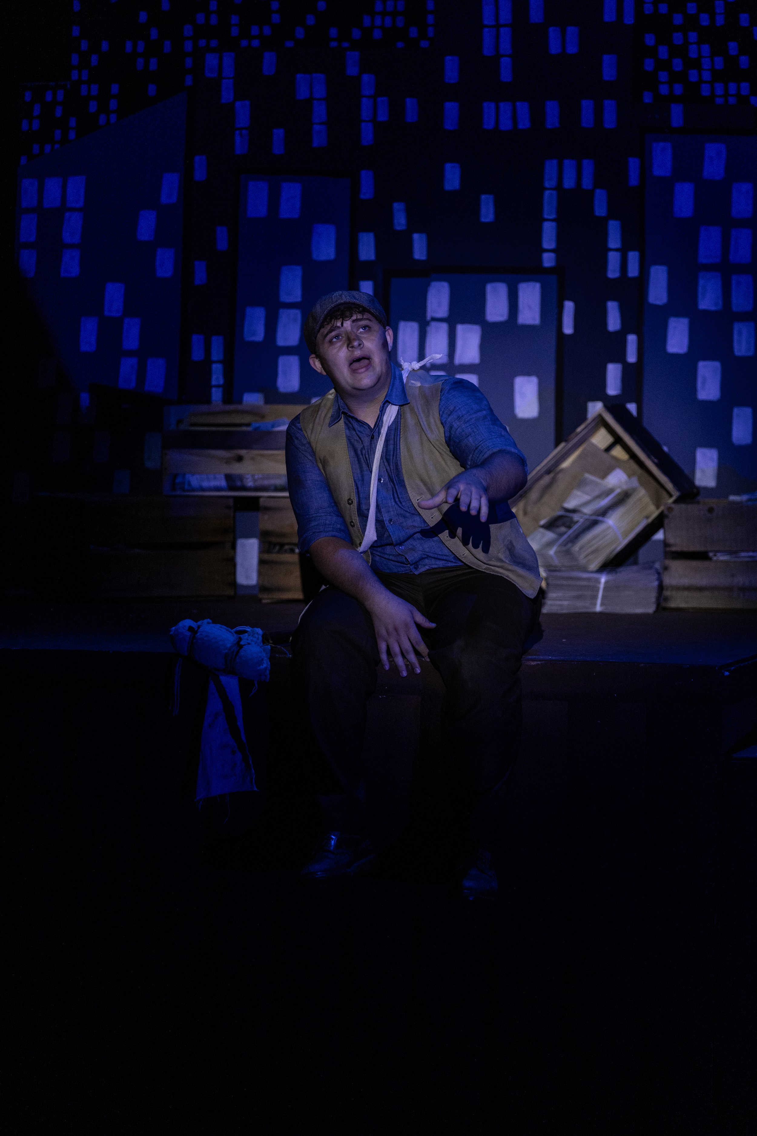 A performer on stage, dressed in a blue shirt and brown vest, sitting and gesturing with his right hand. The background features a stylized cityscape with blue illuminated windows and a dark night sky. There are props and set pieces, including a card