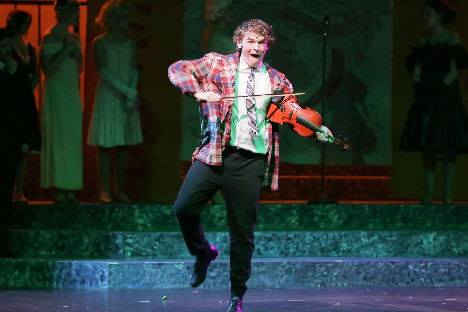 A man in a red plaid blazer and black pants playing a violin on stage, smiling and energetic, with four women in vintage dresses standing in the background.