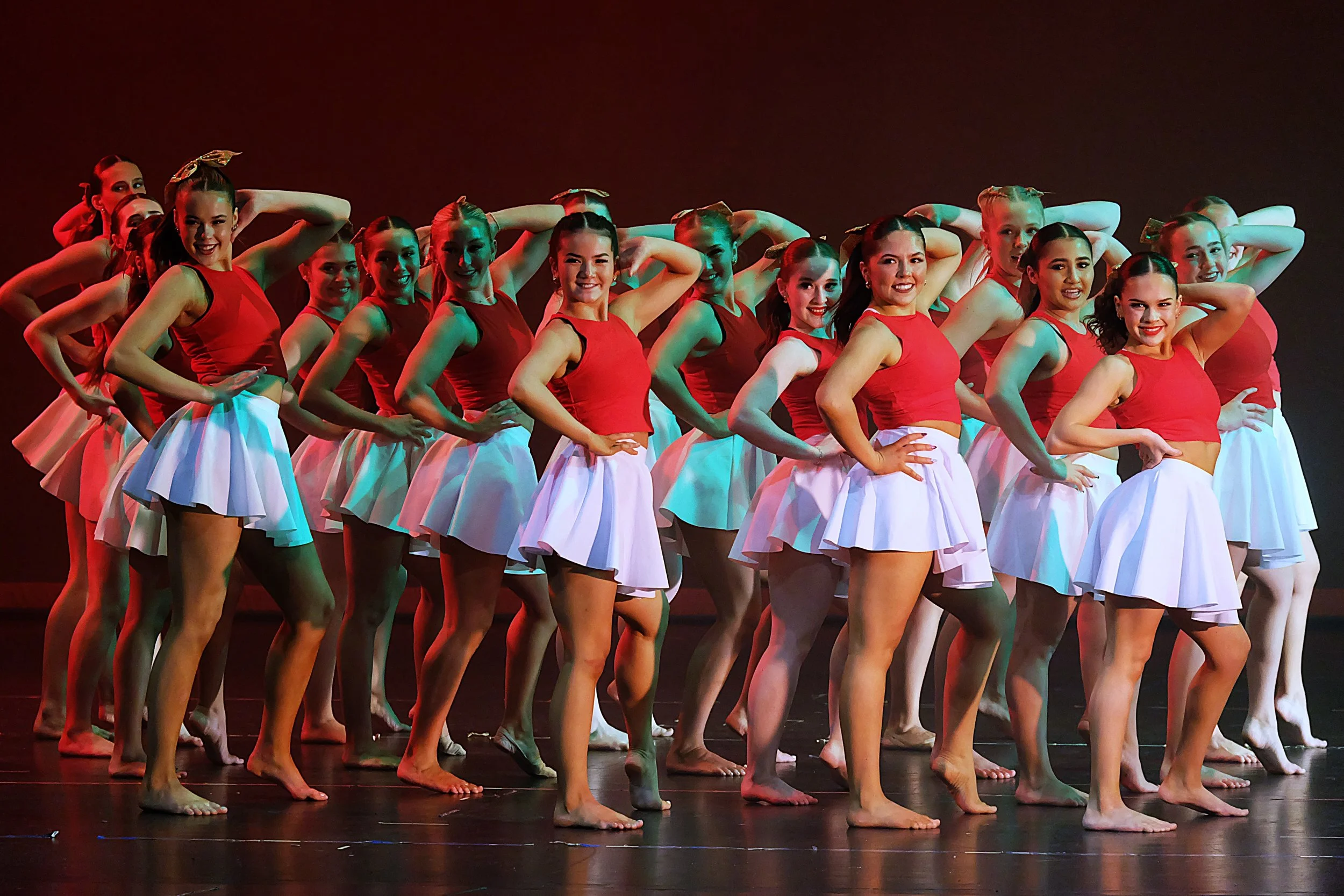 Group of female dancers dressed in red tops and white skirts performing on stage, with some standing and posing with hands on hips, under stage lighting.