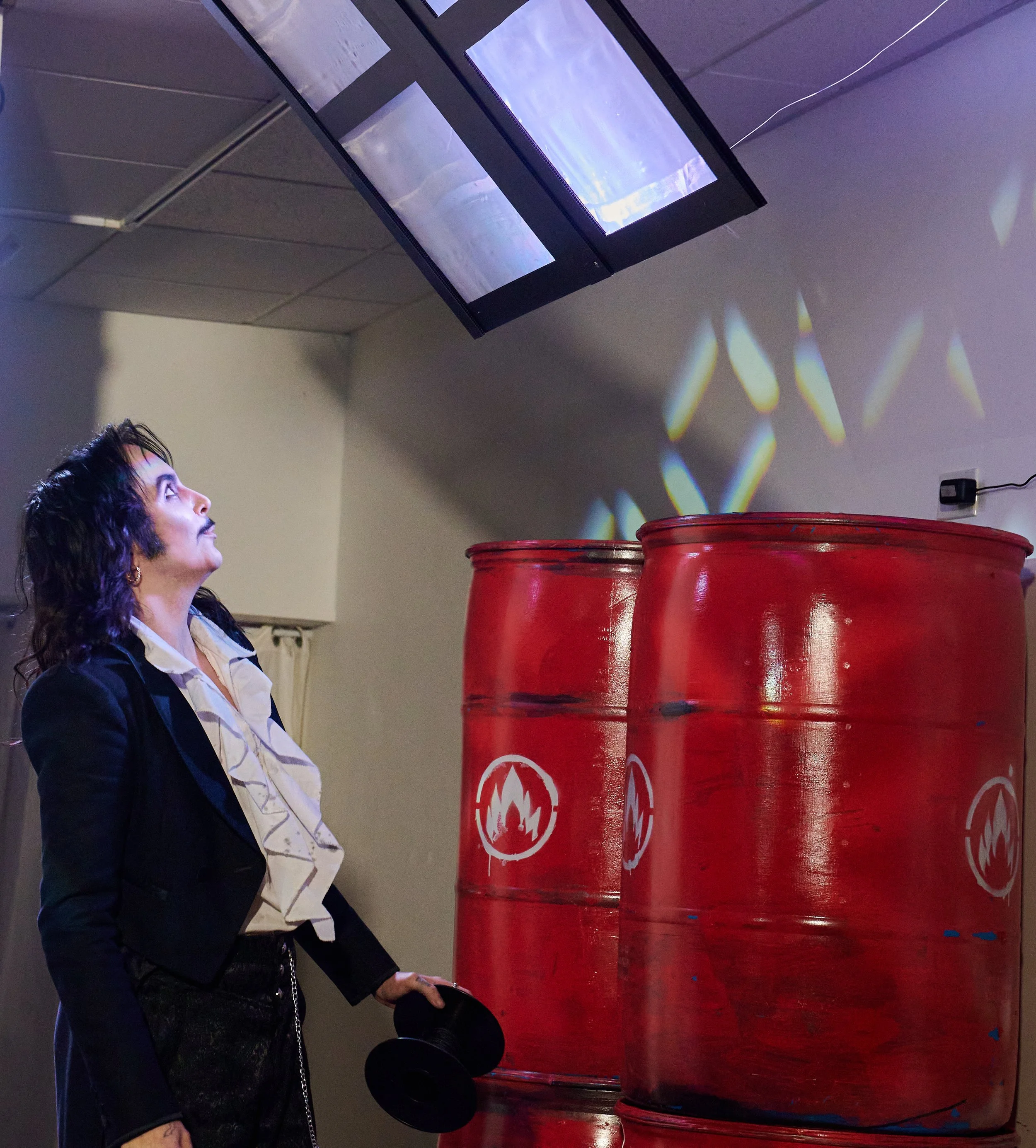 A woman with curly hair looking at a large screen mounted on the ceiling in a room with dark walls. In front of her, there are three large red barrels with some seals or markings on them. The scene is dimly lit, with reflections and patterns dancing 
