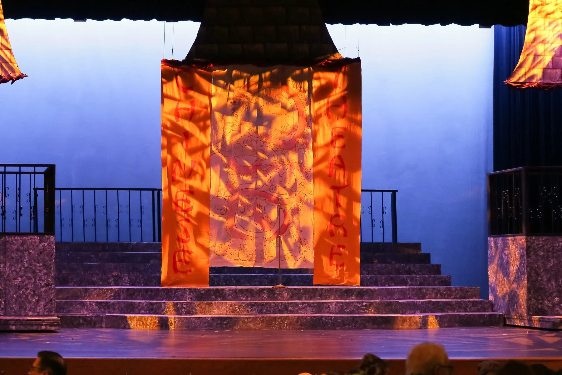 Stage with orange banner featuring a dragon illustration and red text, flanked by black railing and stone stairs, in an auditorium setting.