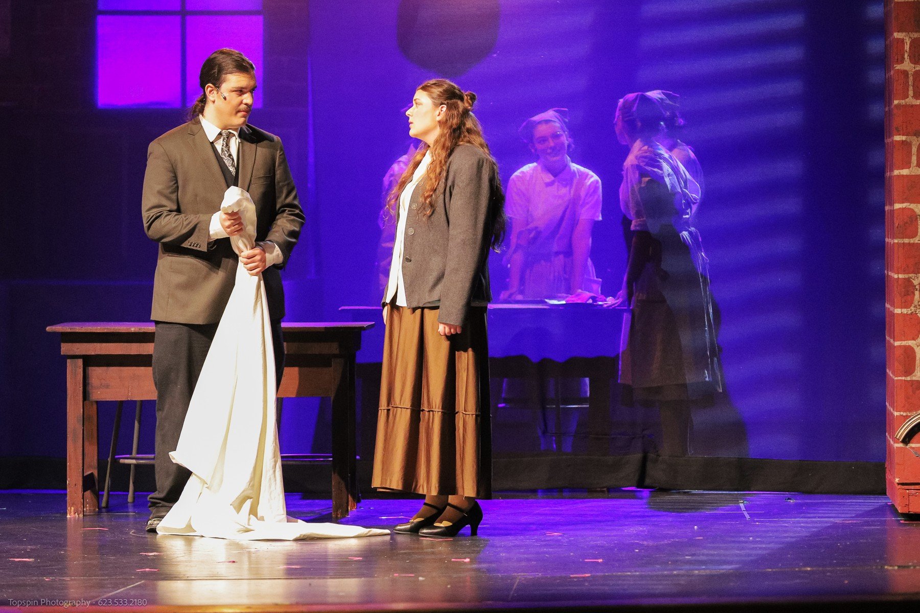 Two actors on stage during a theatrical performance. The man on the left, dressed in a brown suit, holds a white cloth. The woman on the right, wearing a brown skirt and gray blazer, is facing the man. In the background, three women are behind a tran