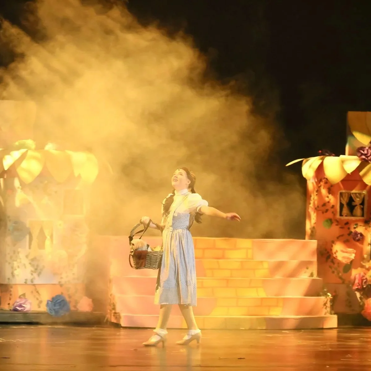 A woman on stage wearing a vintage-style dress, holding a basket, performing under warm stage lights with flowers and whimsical scenery in the background.