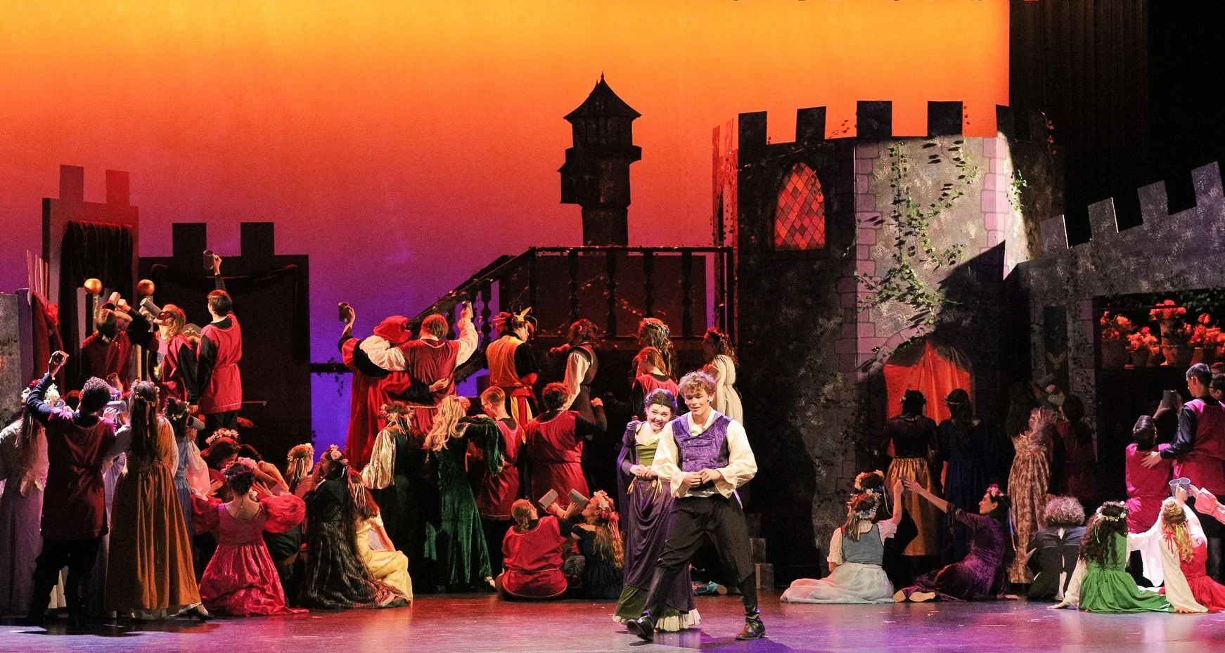 A theatrical stage is filled with actors dressed in medieval costumes, performing a play set in a castle with a colorful sunset backdrop projected behind.