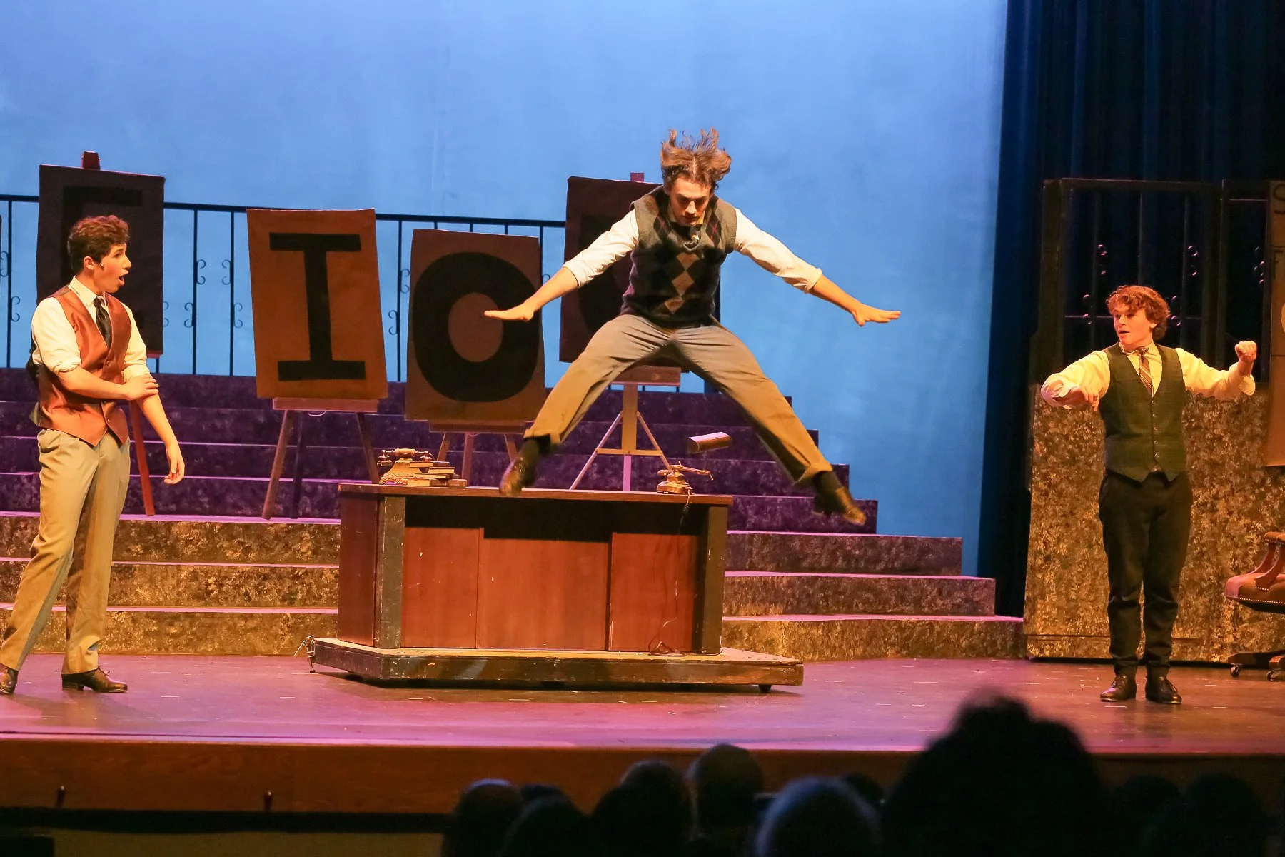 Three young men on stage performing a theatrical play. One in the center is jumping or leaping over a desk, with arms outstretched and hair flying. The other two young men are standing on either side, one on the left with a surprised expression and t