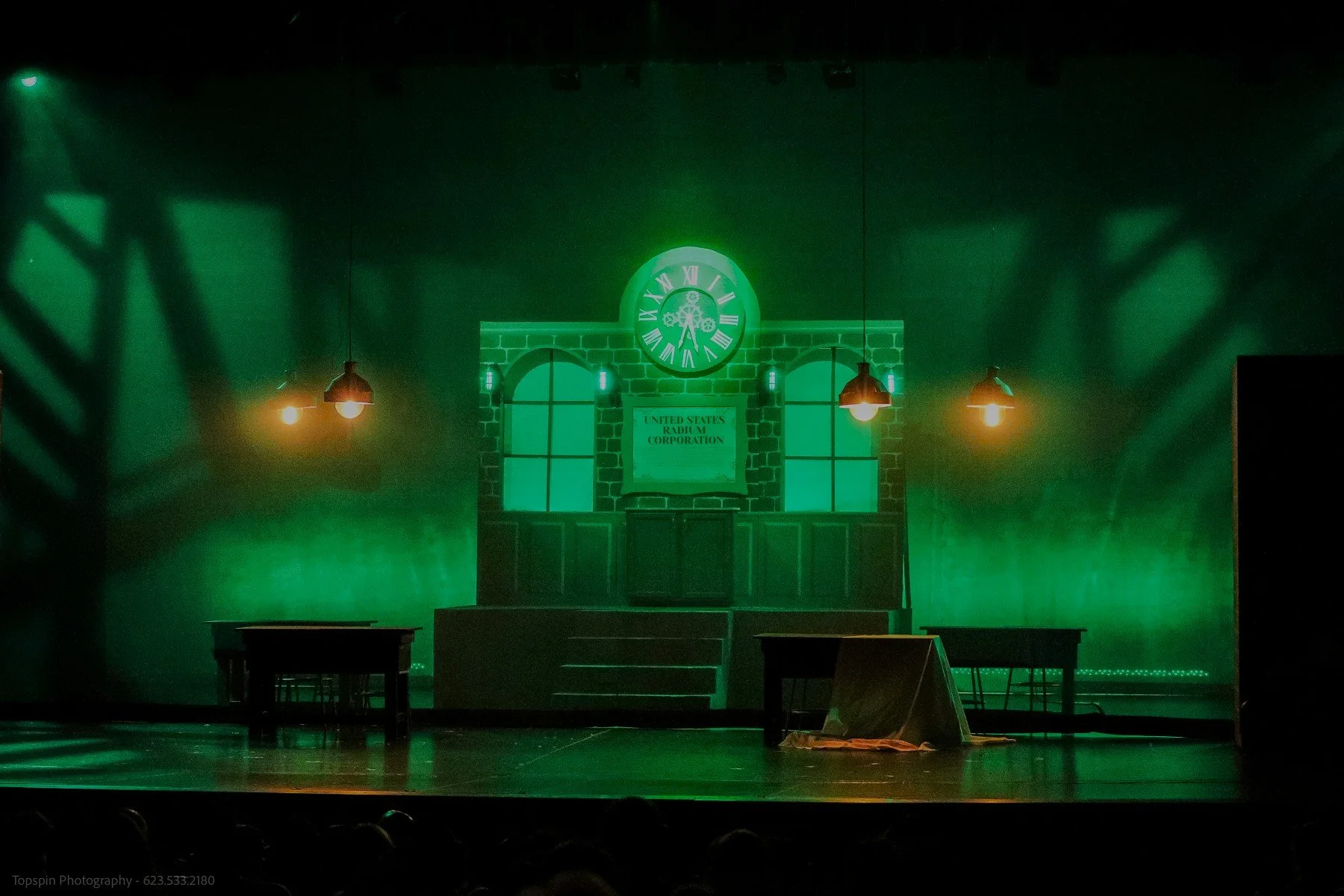 The stage has a green light with a backdrop resembling a building with two windows, a clock showing almost 8:00, and a sign that reads 'United States Radio and TV Corporation'. There are hanging lamps illuminating the scene and a few tables and chair