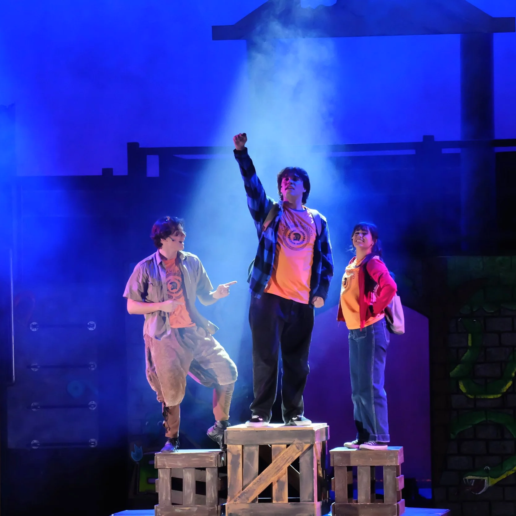 Three young people perform on stage with theatrical lighting. One person stands on a wooden crate with their fist raised. The others are smiling and standing on the platform, all wearing casual clothes.