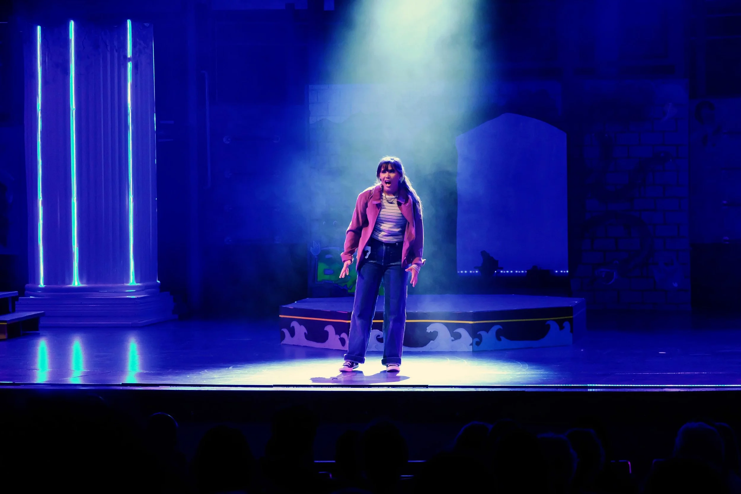 A young woman with dark hair and bangs performing on stage, wearing a striped shirt, a pink jacket, and jeans, under dramatic blue lighting with a misty effect in the background.