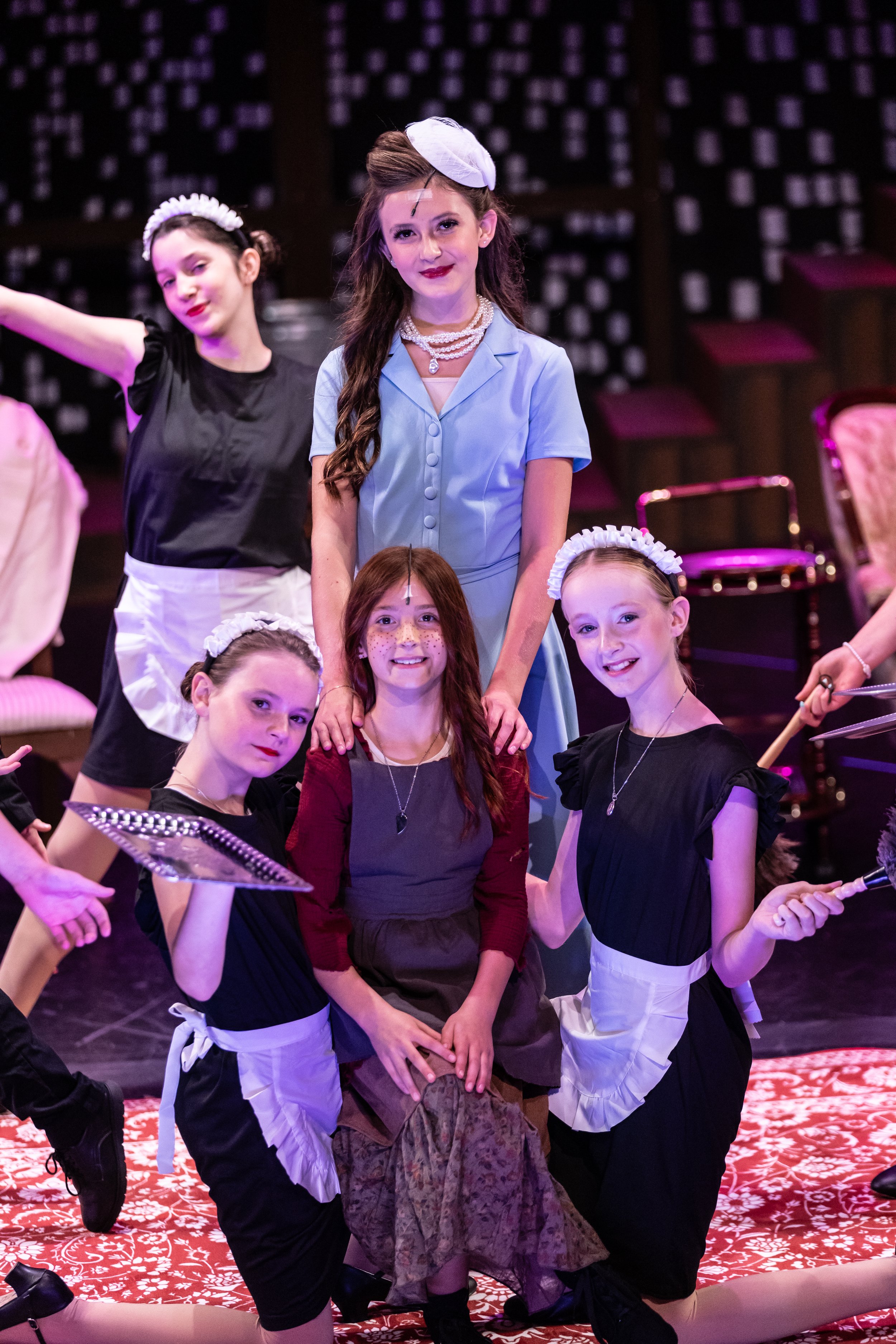 Women and girls dressed in costumes on stage, some in aprons and headbands, performing in a theatrical production or play.