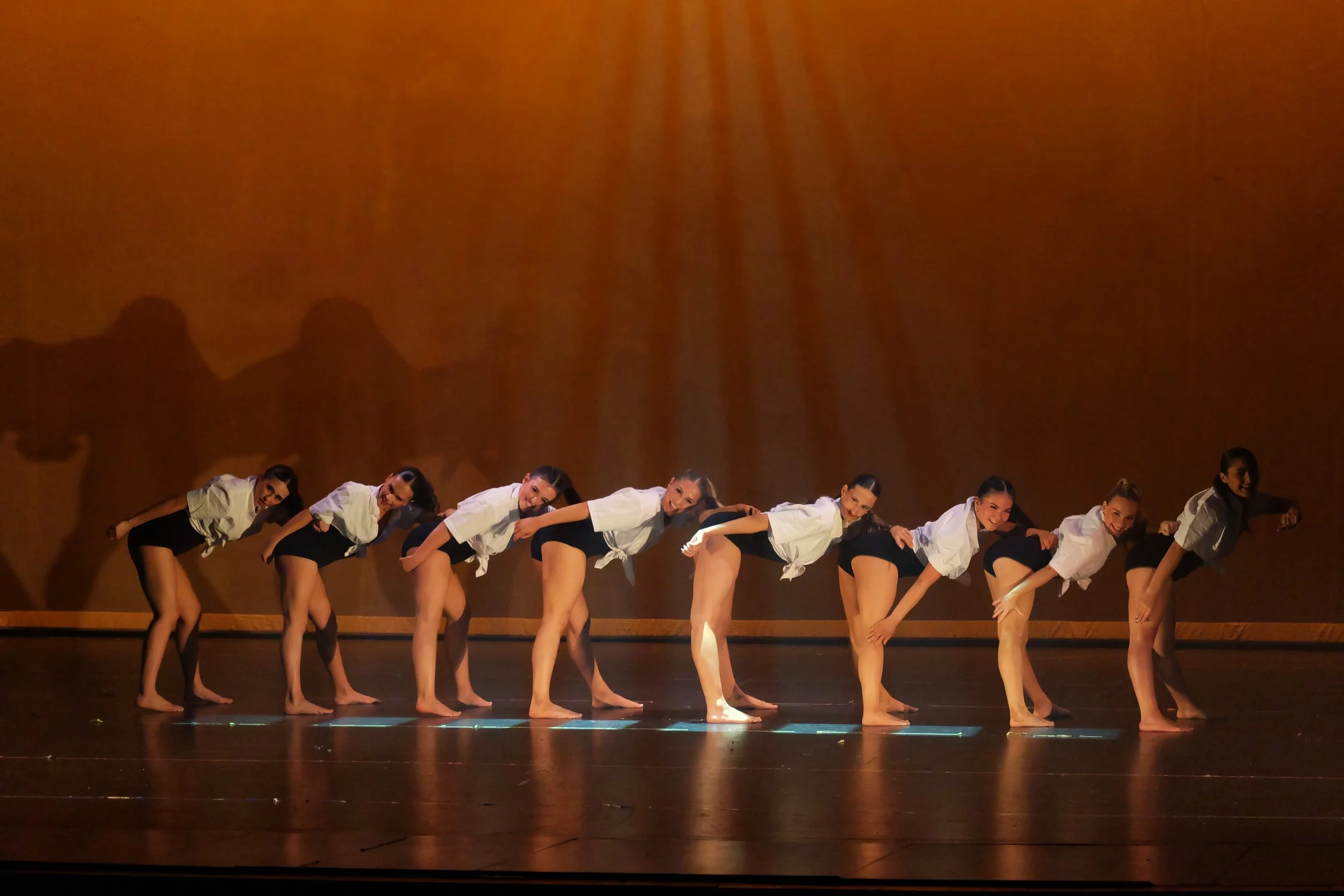 A group of dancers in white shirts and black shorts performing a synchronized dance move on stage with a brown backdrop and their shadows cast on the wall.