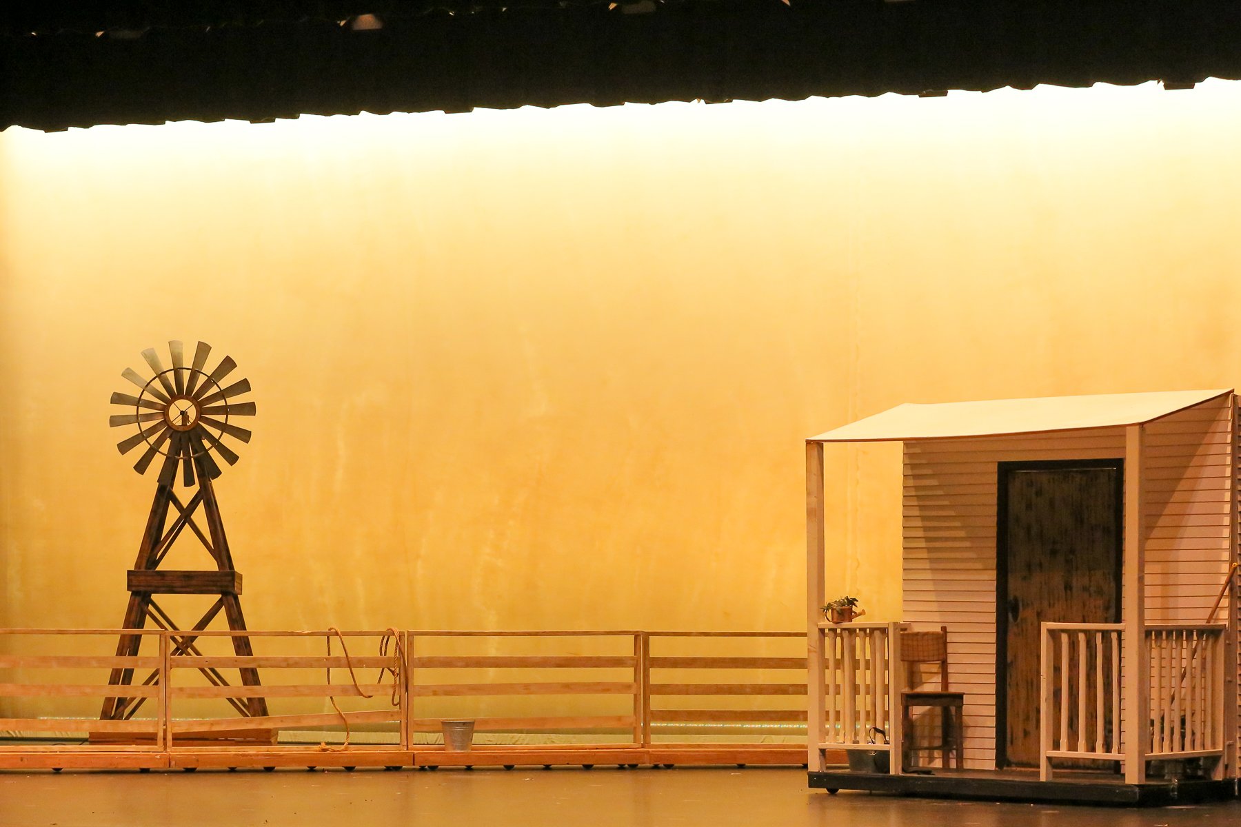 Stage with a windmill and a small house, surrounded by a wooden railing, set against a plain yellow backdrop