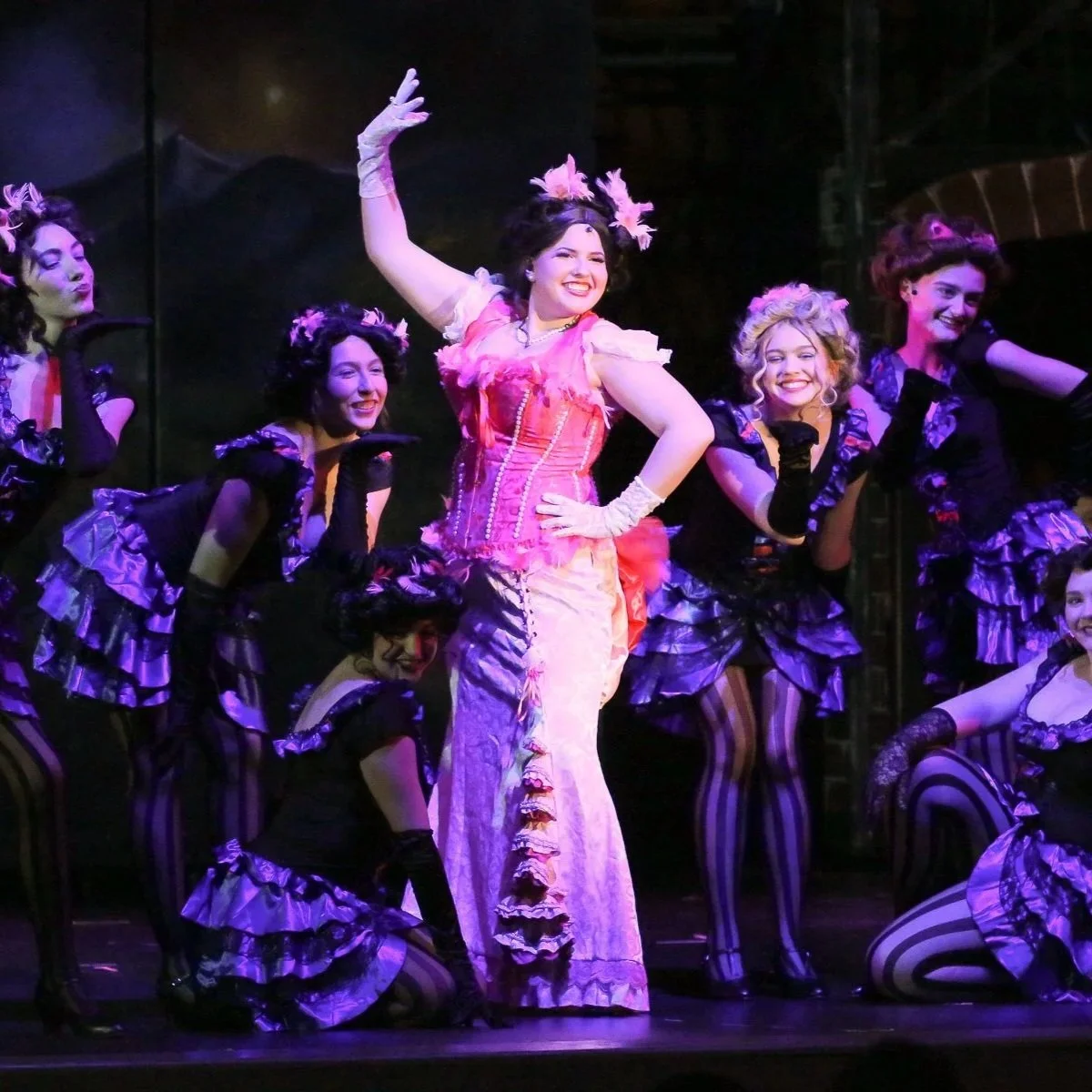 Theater cast of women in colorful costumes, with one woman center stage in a pink dress, waving, surrounded by others in black and purple, performing on stage.