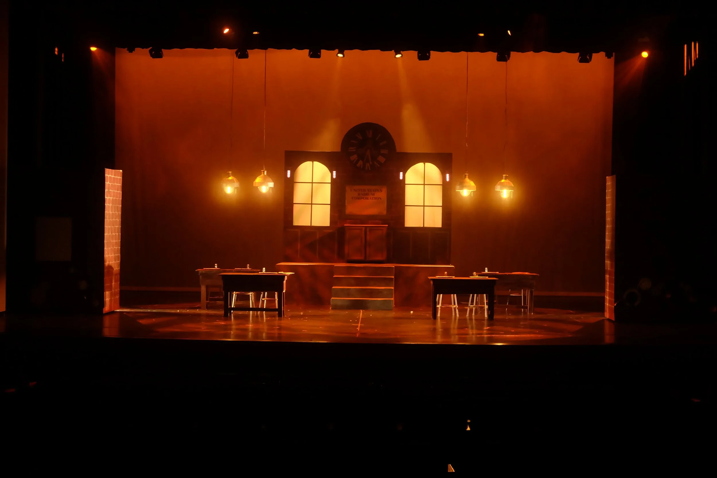 A theater stage set for a play with warm lighting, a backdrop resembling a sunset sky, large windows, a central clock, and furniture like tables and stools.