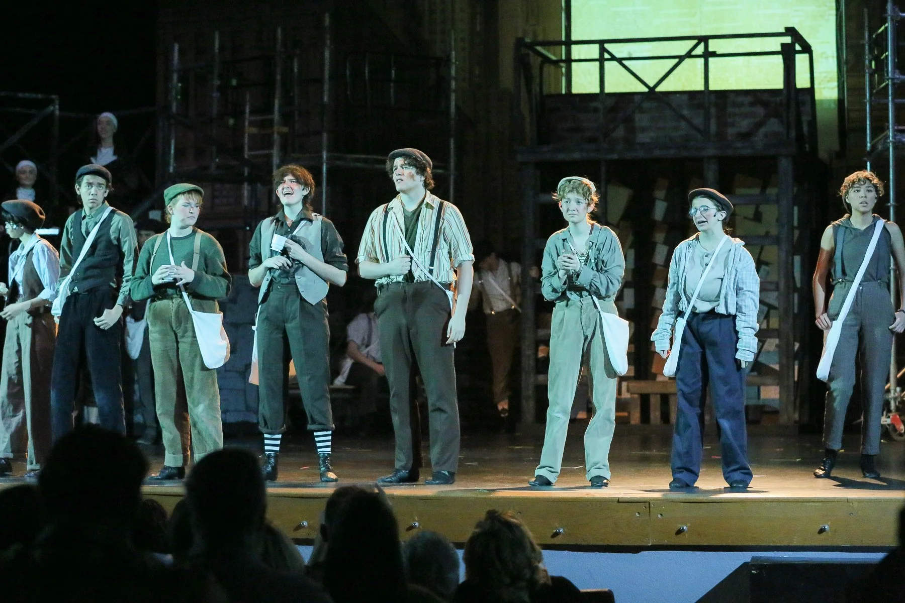 A group of young actors on stage performing in a play with a theatrical set background and audience visible at the front.