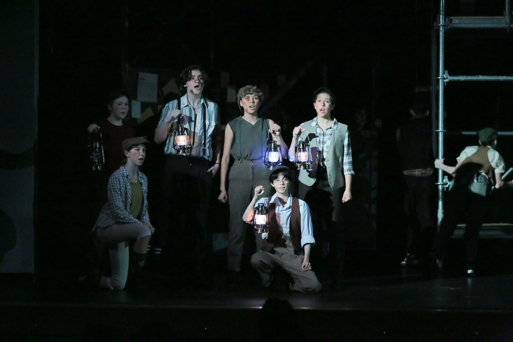 Group of children on stage performing in a play, each holding a lantern, with a dark background and some stage scaffolding