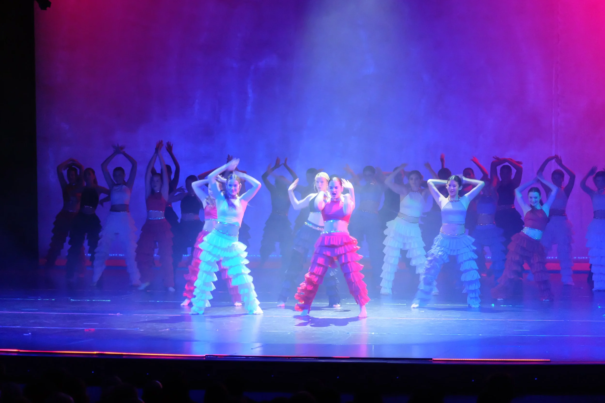 Group of dancers performing on stage, with some wearing white and pink costumes with ruffled skirts, and others in dark costumes, under purple and blue stage lighting.