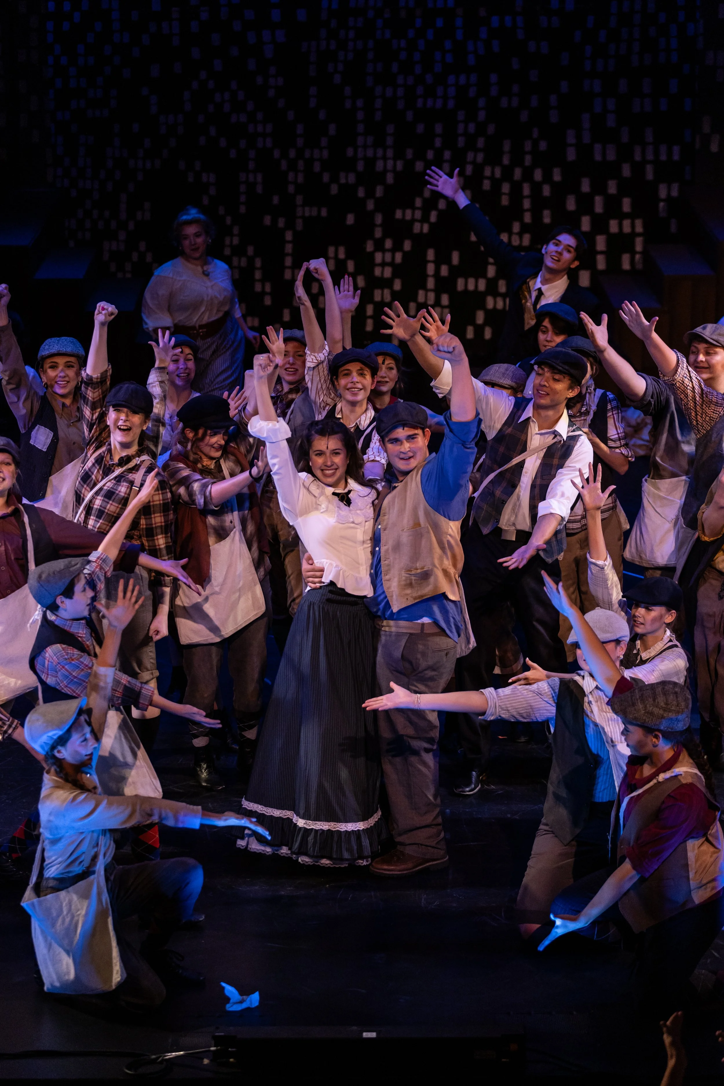 A theatrical stage scene with a diverse group of young performers dressed in vintage, early 20th-century clothing, including caps, plaid shirts, and vests. They are smiling and raising their hands in a lively, celebratory manner. The background featu