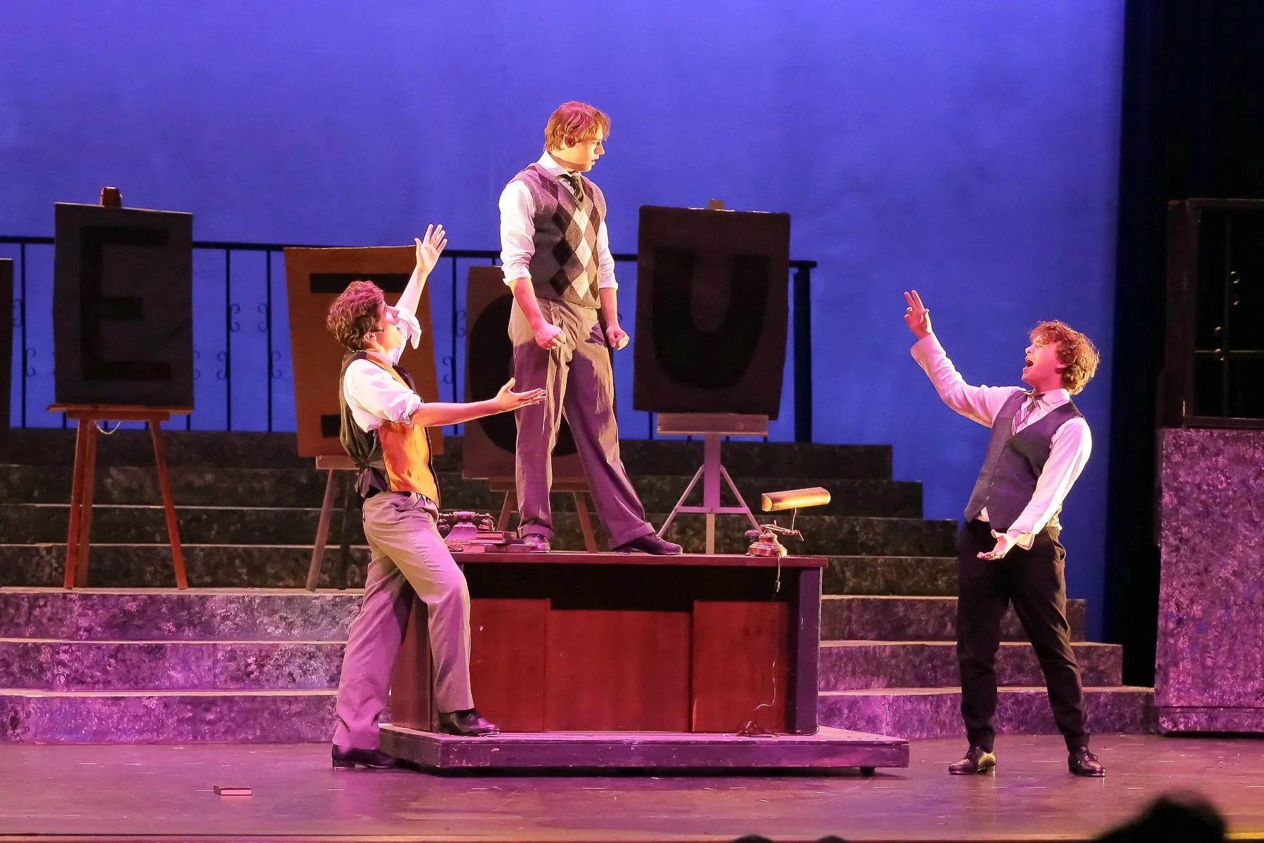 Three young boys with curly hair performing on a stage, dressed in vintage-style clothing, with one standing on a platform and two on the stage, engaging in a theatrical scene with expressive gestures, set against a dark blue background with stage pr