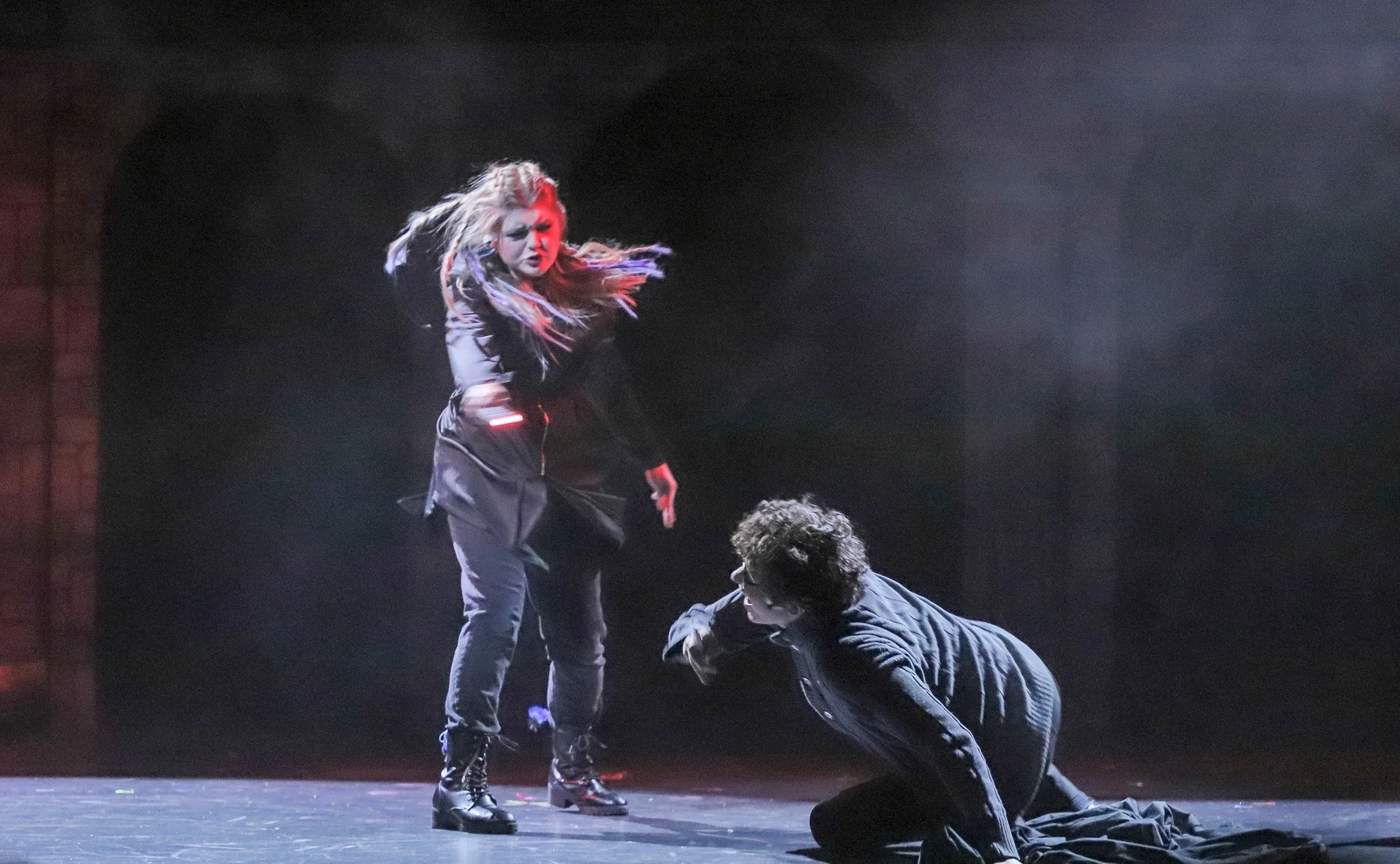 A woman with colorful braided hair and dark clothing appears angry as she confronts a man on the floor during a theatrical performance. The scene is lit dramatically with smoke effects.