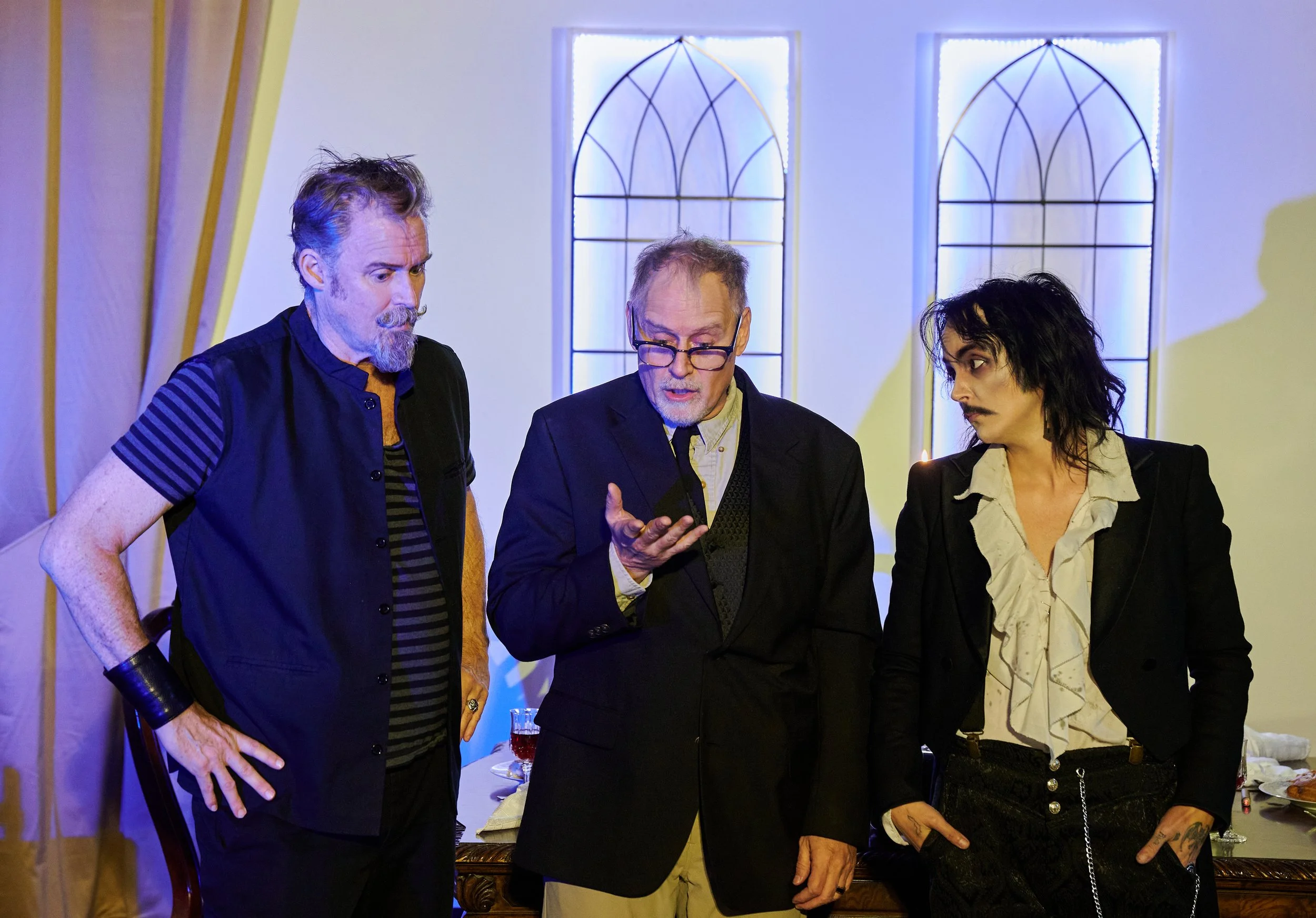 Three people are engaged in a conversation at a gathering in a room with decorative stained glass windows. One man with glasses is speaking while the others listen.