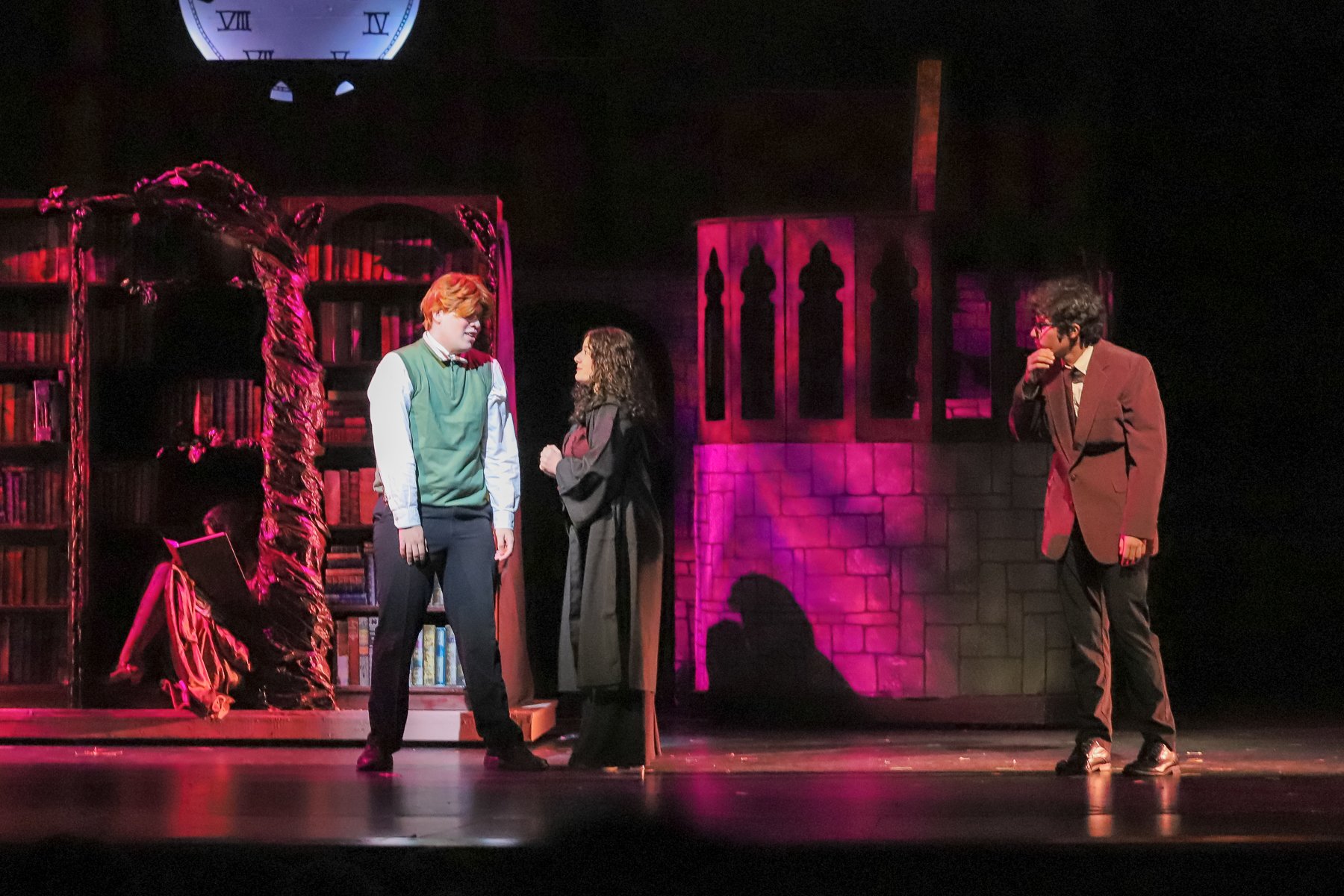 Three actors on stage performing a scene in a play. The stage set includes bookshelves, a stone wall backdrop, and purple lighting. The actor on the left has red hair, is wearing a green and white outfit, and is engaged in conversation with a woman i