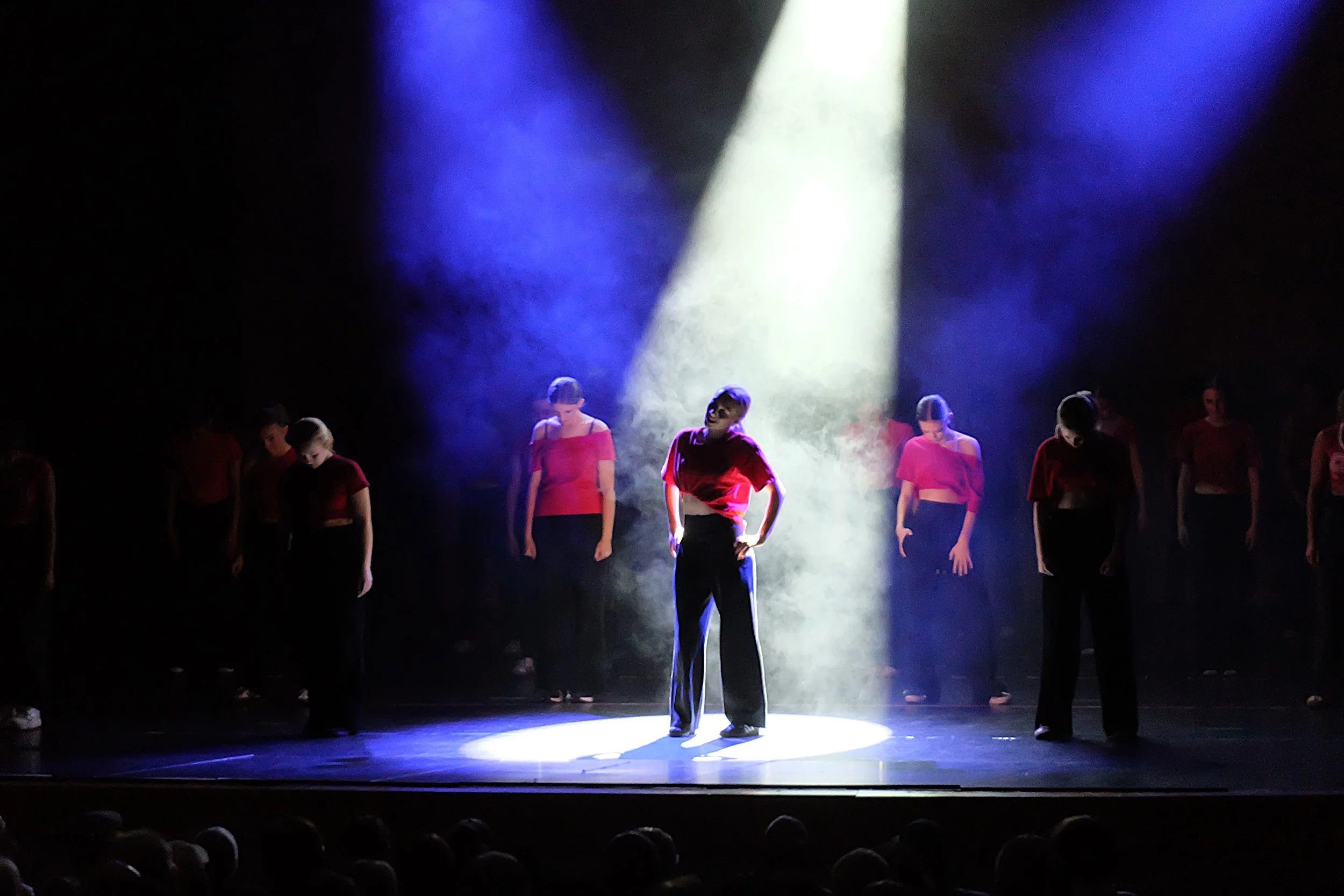 A group of dancers on stage, illuminated by a bright white spotlight, wearing matching red tops and black bottoms, performing a choreographed dance routine in front of a dark background.