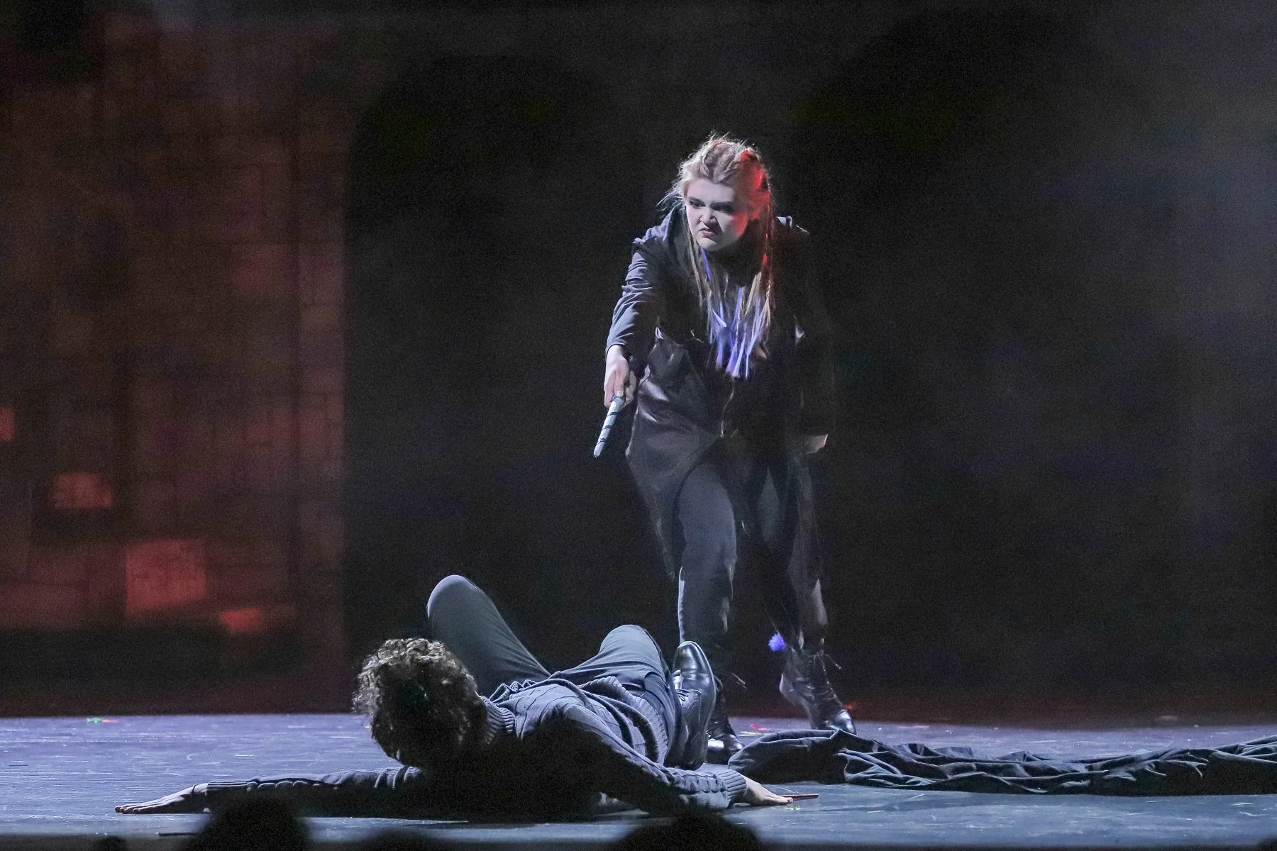 A theatrical scene with a woman standing over a man lying on the stage floor. The woman looks angry or intense, holding a microphone in one hand. The stage is dark with minimal background detail.