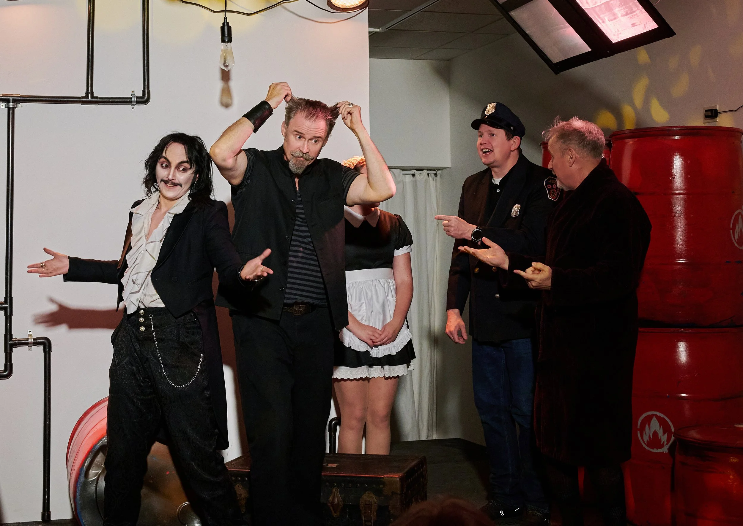 People dressed in costumes, possibly for a theatrical or themed event, standing on a stage in a room with industrial pipes, light bulbs, and a large red barrel.