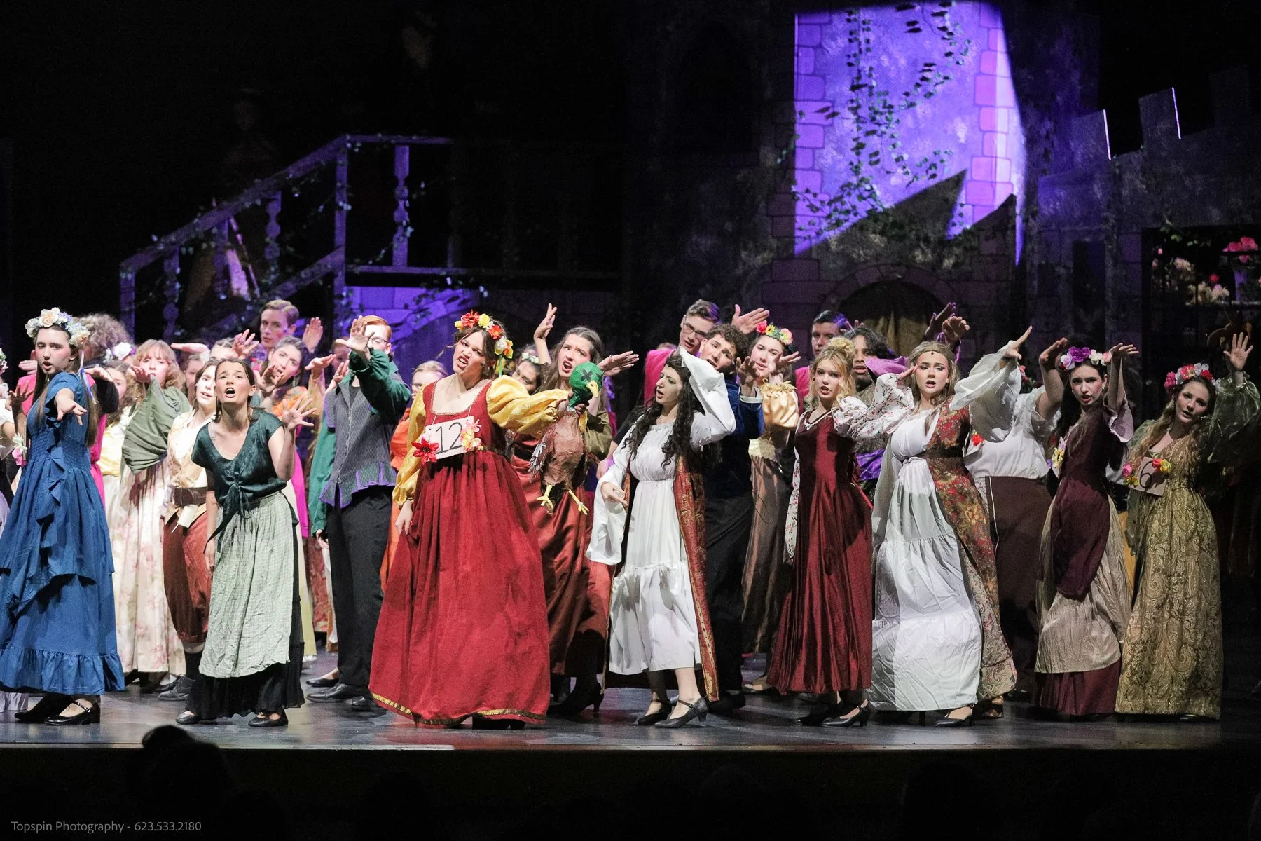 A group of performers on stage in colorful period costumes, singing and dancing, during a theatrical production with a dark background and purple lighting.
