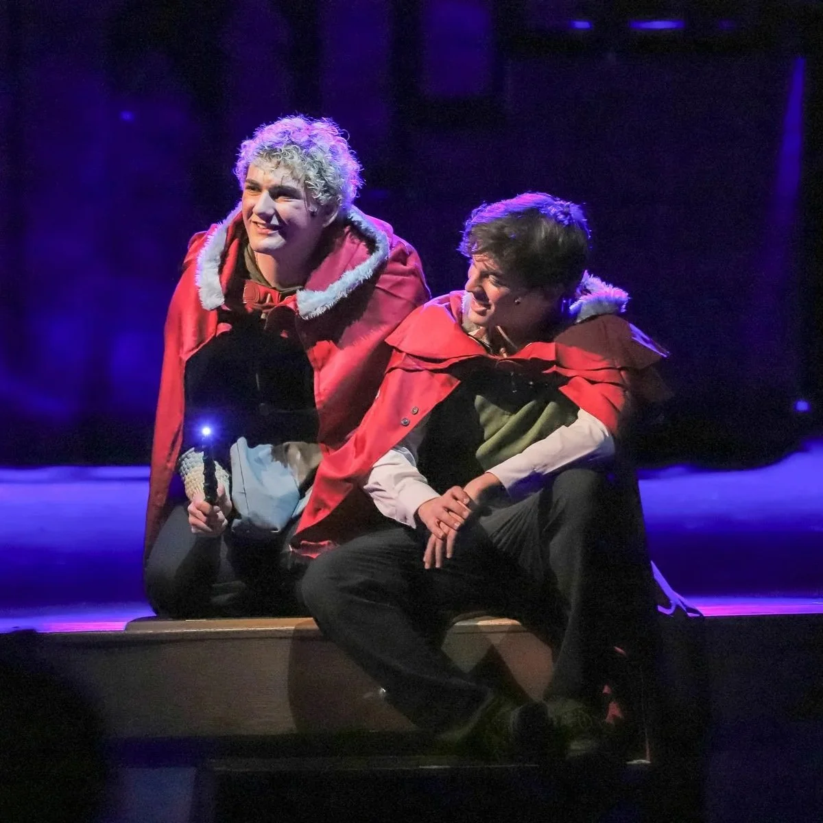Two actors on stage dressed in red capes, one with curly blond hair and the other with dark hair, performing in a theatrical production with dramatic purple lighting.