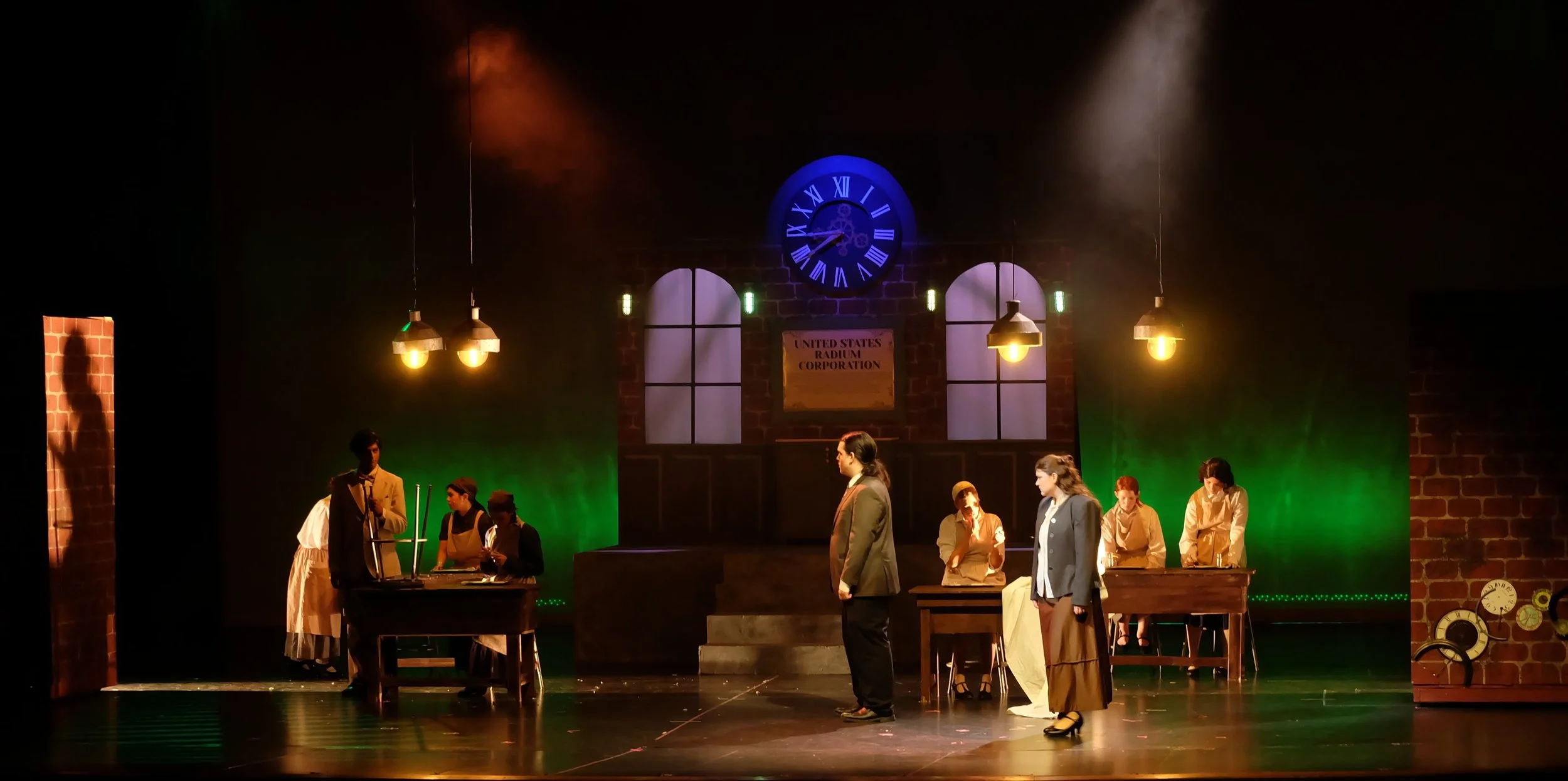 A theatrical stage set mimicking an office scene with actors in period costumes, a large clock with Roman numerals, brick walls, and hanging lamps creating a warm ambiance.