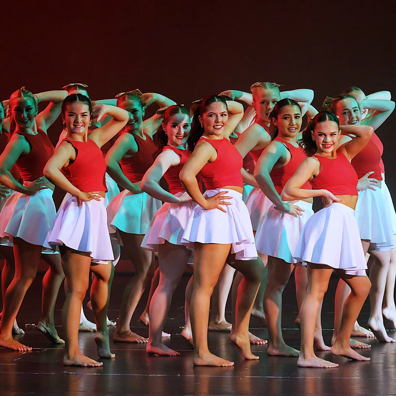 A group of young women dressed in red sleeveless tops and white skirts performing a dance routine on stage. They are standing with hands on hips, smiling, with some dancers in the background. The stage is dimly lit with a dark background.