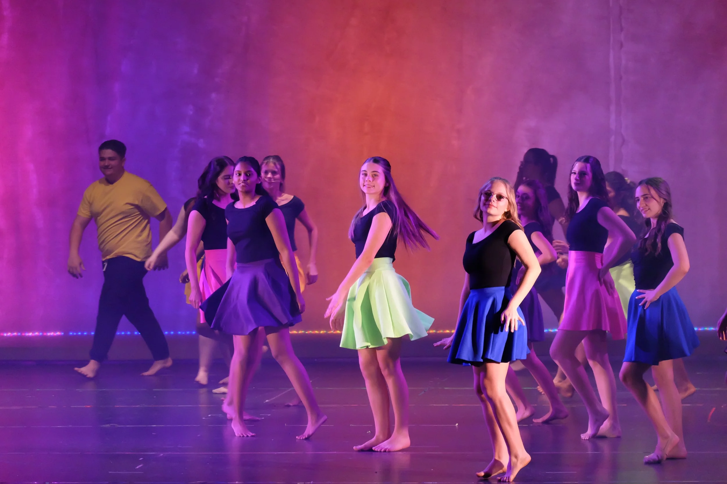A group of female dancers on stage in black top and colorful skirts, with purple lighting in the background.