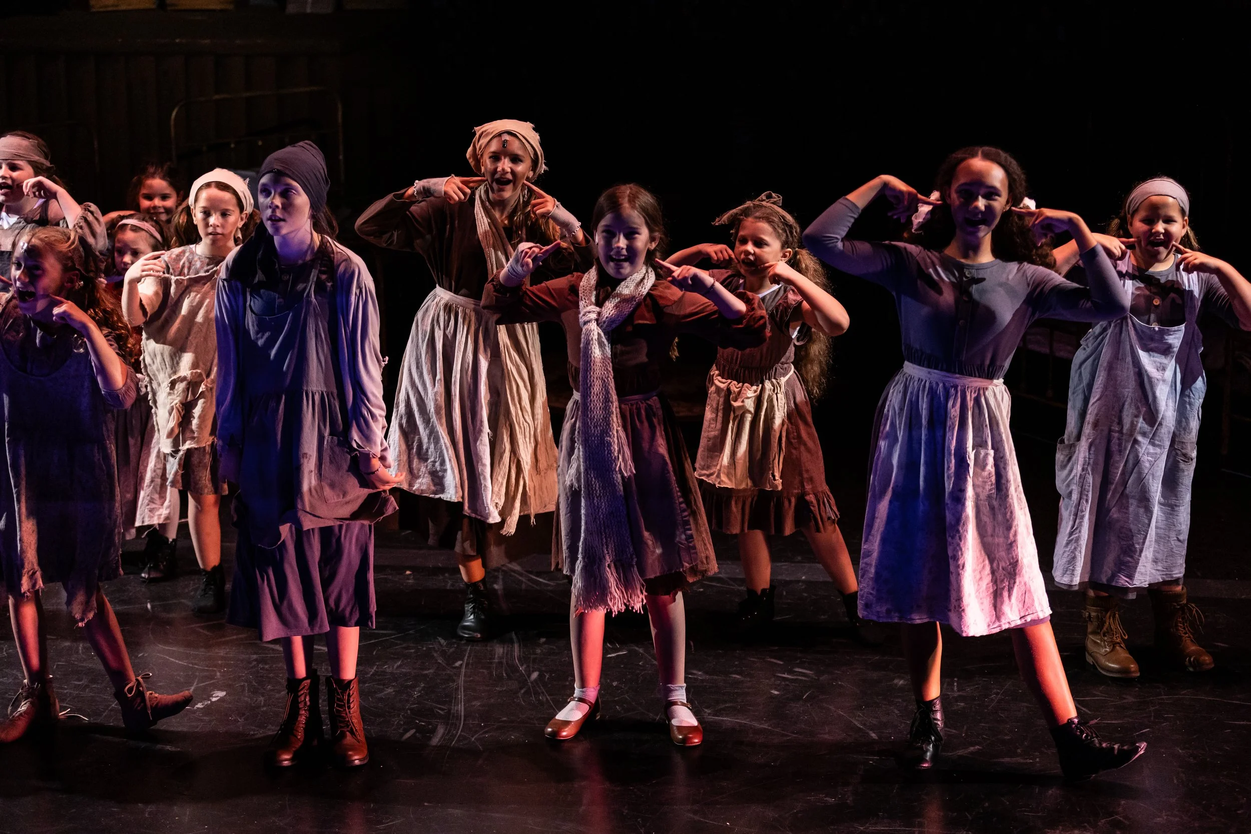 Group of children performing on stage in vintage costumes, striking a pose with fingers near their heads.