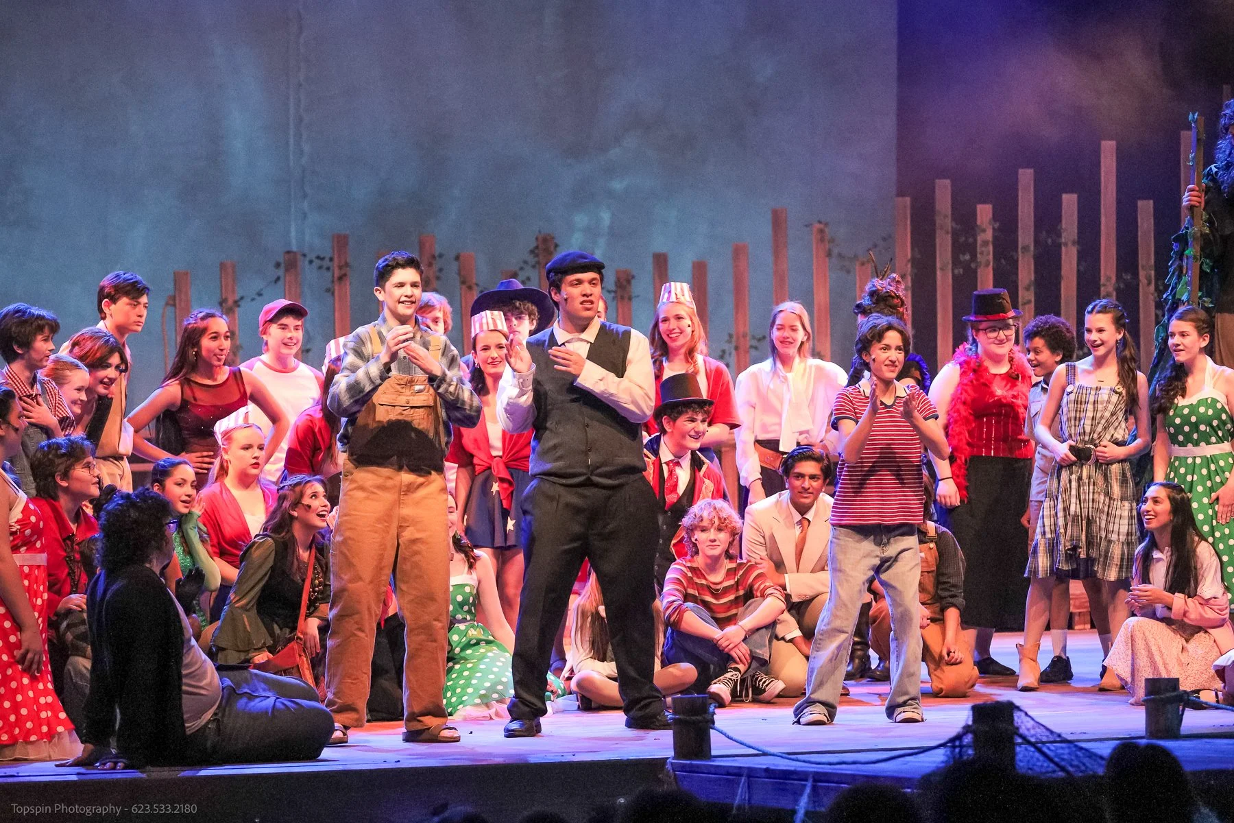 A large group of children on stage during a theatrical performance, dressed in colorful costumes and performing for an audience.