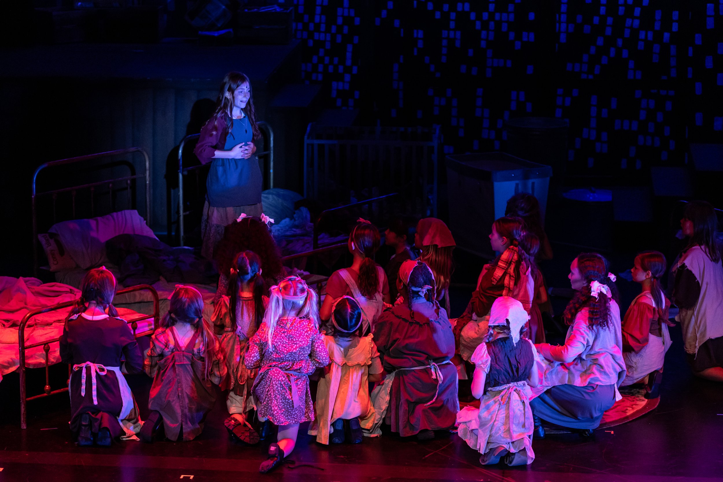 A young girl dressed as a character from a play or musical, standing on stage with her hands clasped in front of her. She is surrounded by children sitting on the floor in costumes, watching her perform. The background features a dark stage with a bl
