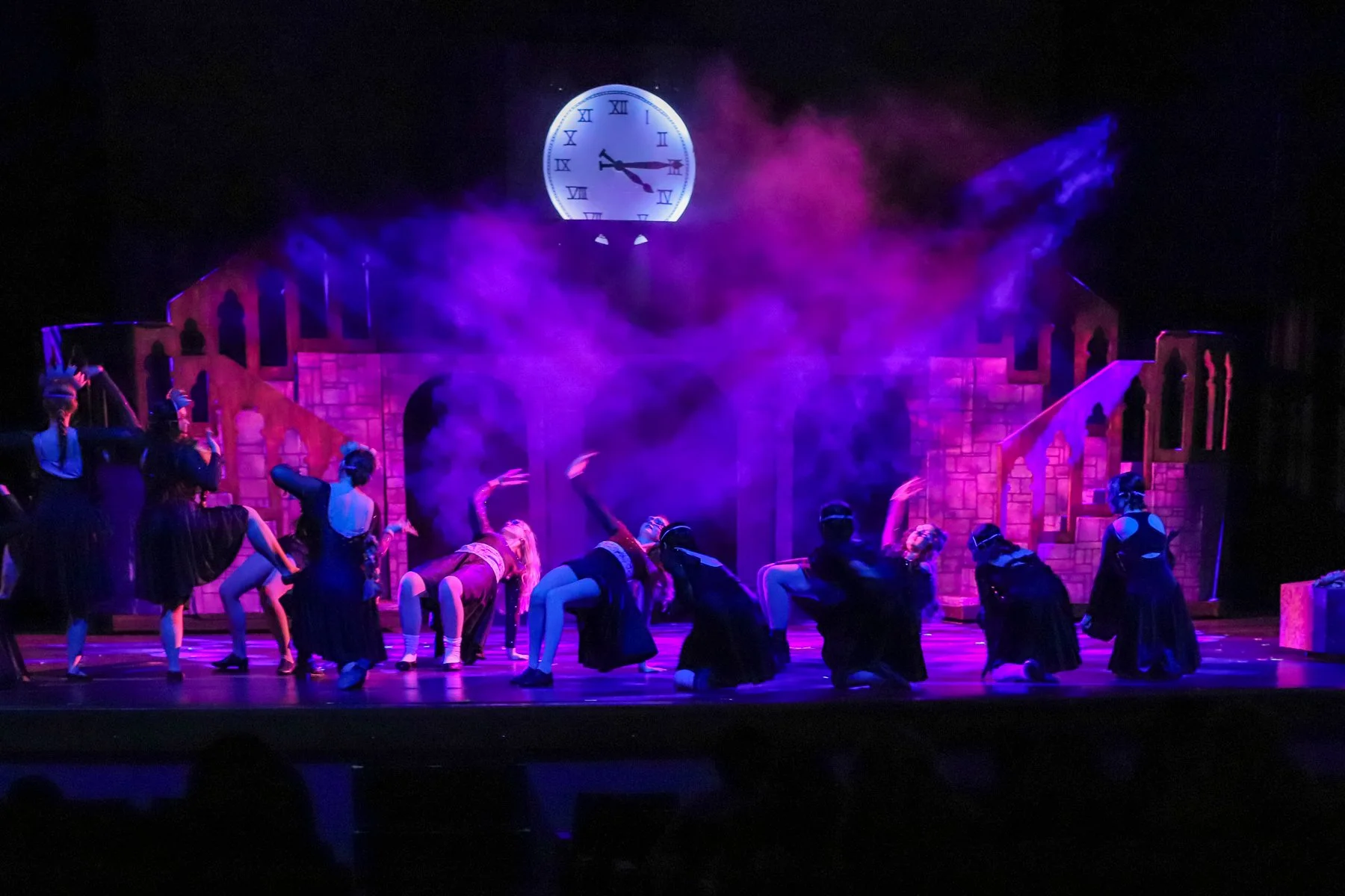 A theatrical stage with dancers in dark costumes performing in front of a large illuminated clock face. The stage is decorated like a castle with brick walls and stairs, and colorful smoke effects are present, with purple and pink lighting creating a