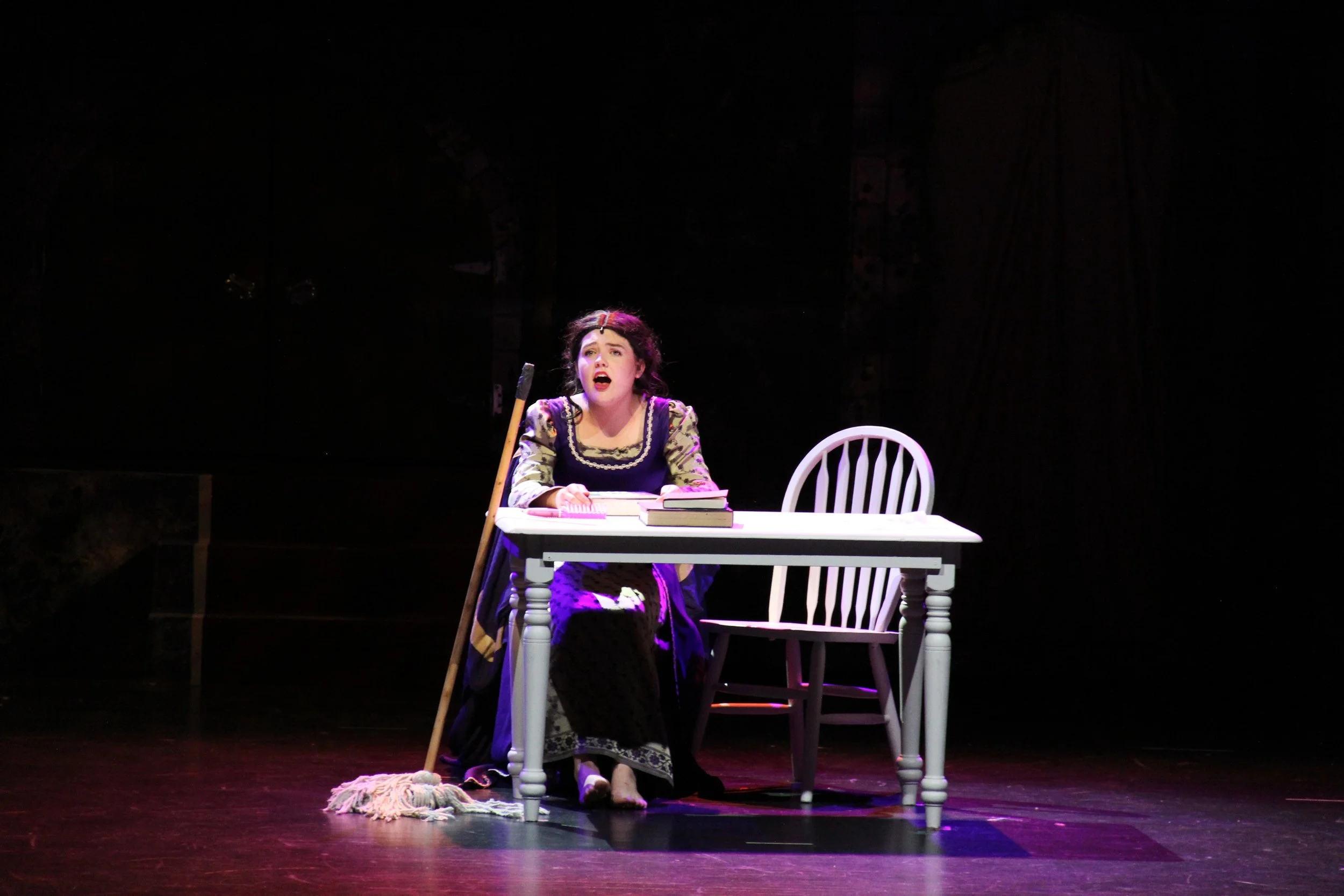 A young woman with curly hair dressed in period costume sitting at a white table on stage, looking distressed, with books and a mop on the floor nearby.