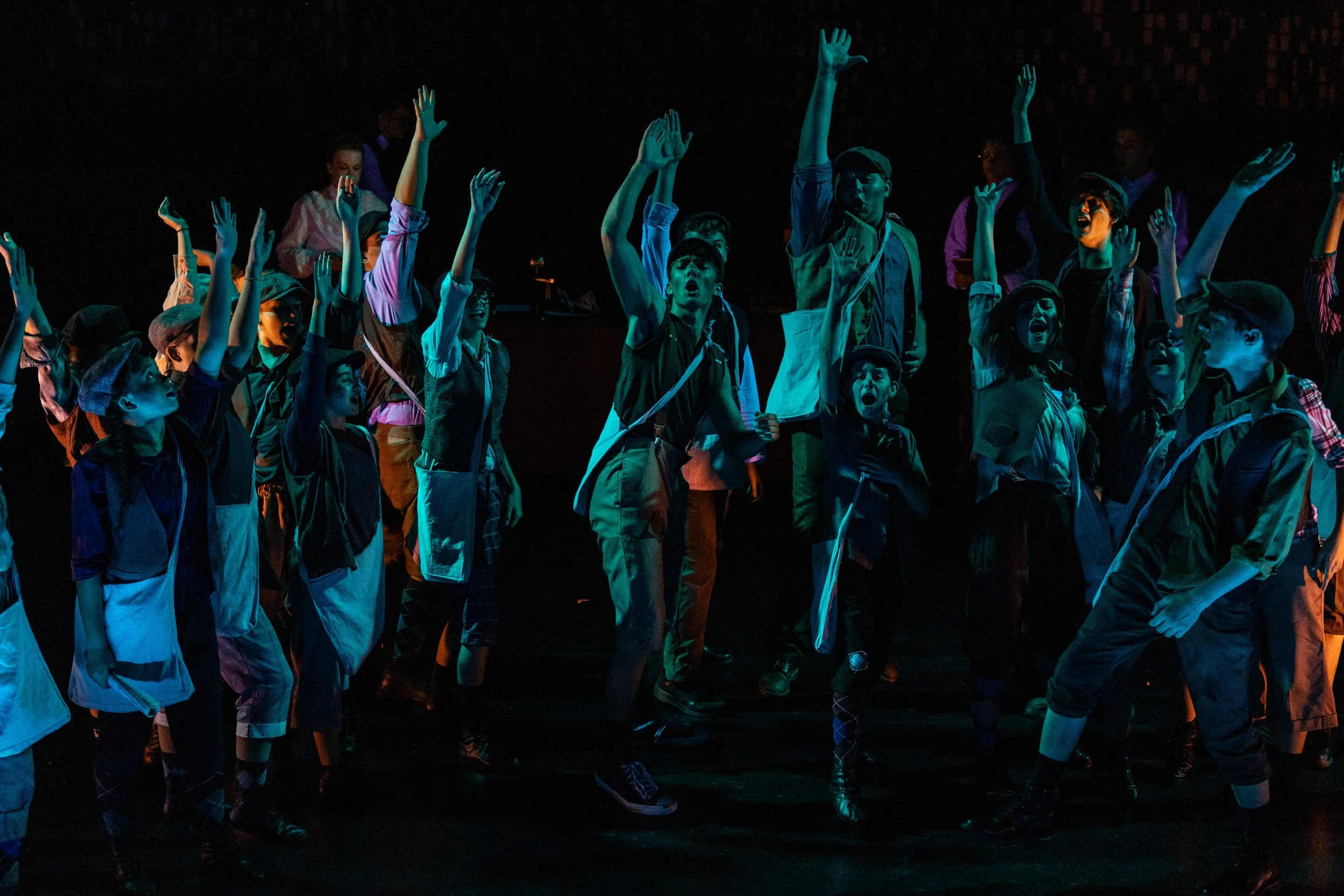 A group of young children dressed in costumes and vests, standing closely together on a dark stage with their arms raised, singing or performing in a play.