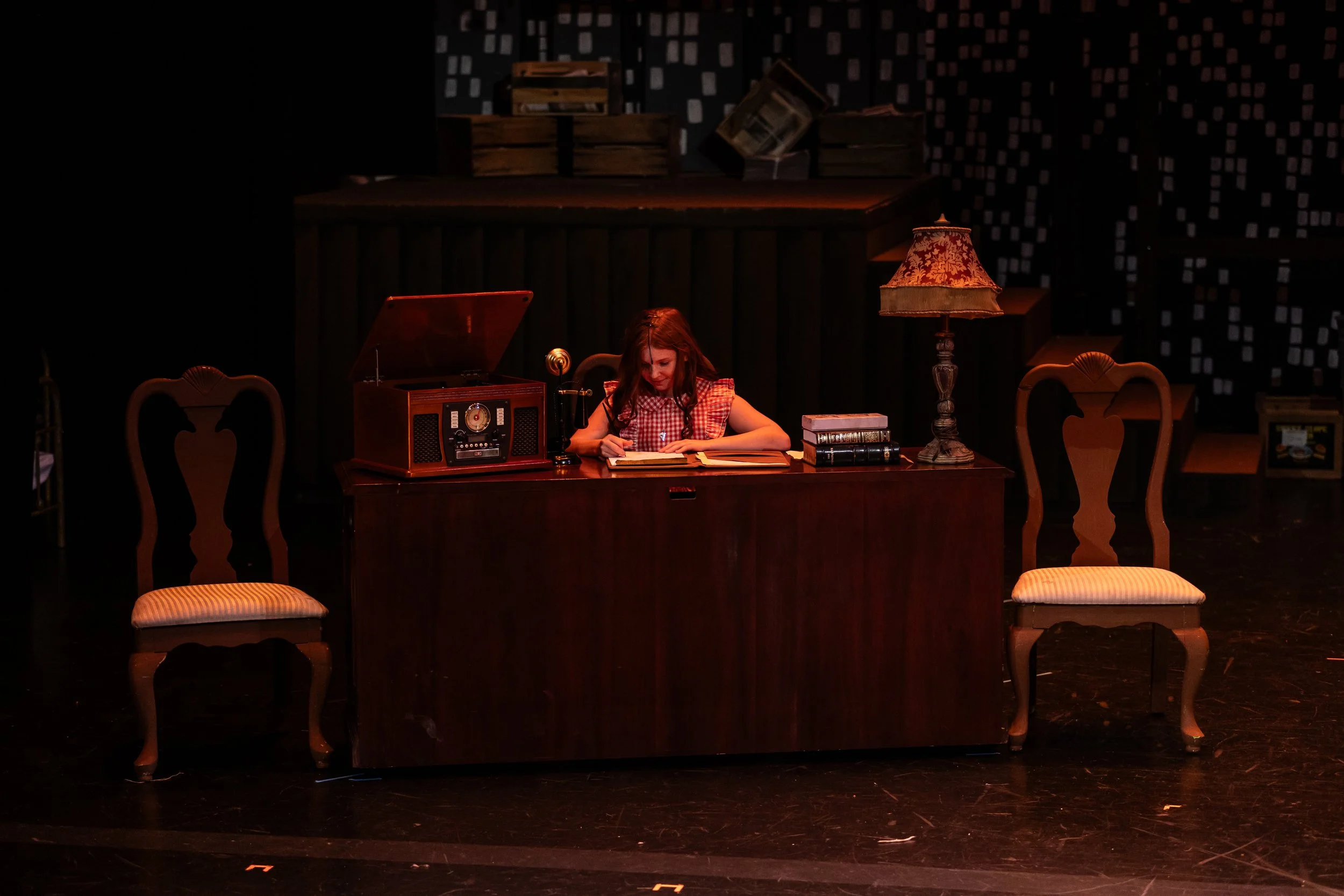 A young girl with red hair wearing a red checkered dress is sitting at a wooden desk on a stage, reading a book. The desk has a vintage radio, a microphone, a stack of books, and a vintage lamp. The background is dark with a patterned design, and the