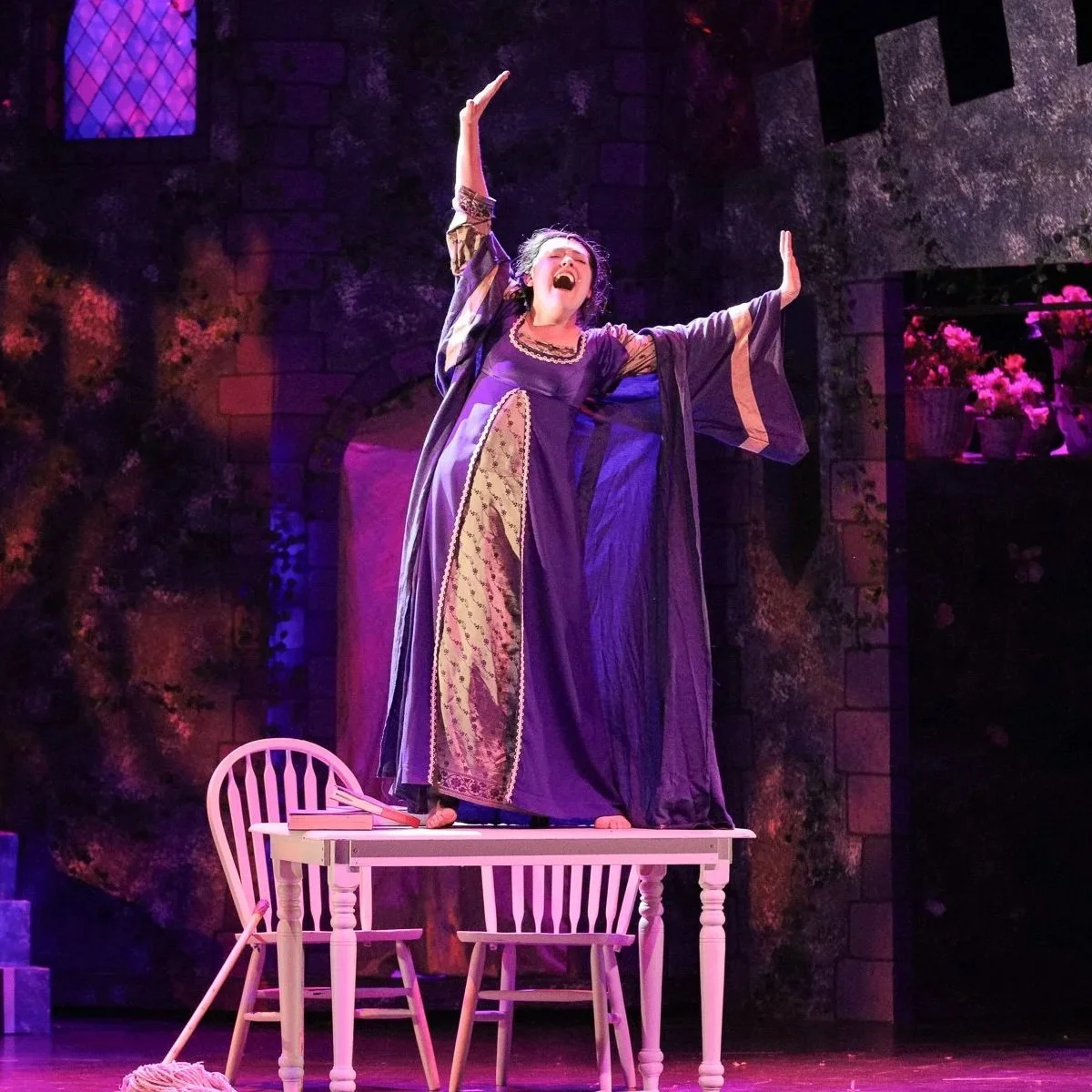 A woman in a purple and gold gown performing on a stage, standing with arms raised on a table. The background features a stone wall and window with pink flowers nearby.