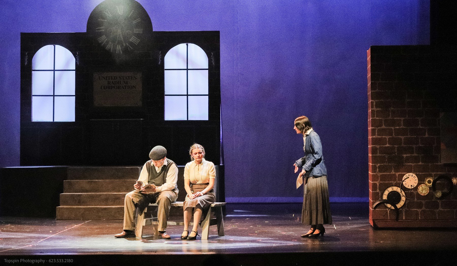 A theatrical stage scene with four actors, three seated on a bench and one standing, in front of a set designed as a building with a clock and windows, with a purple backdrop.