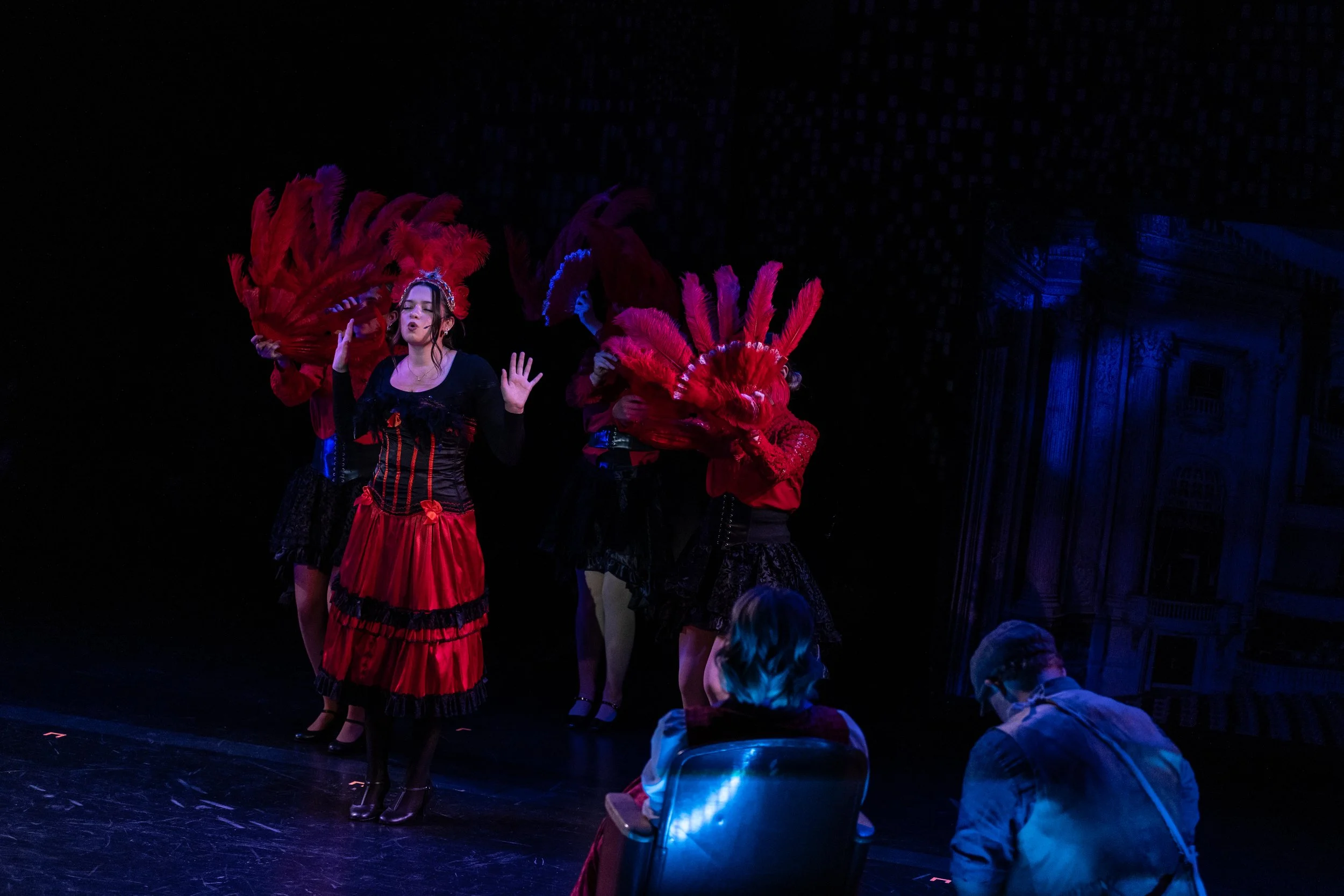 Performers in colorful costumes with large red feathered headdresses on stage during a show.