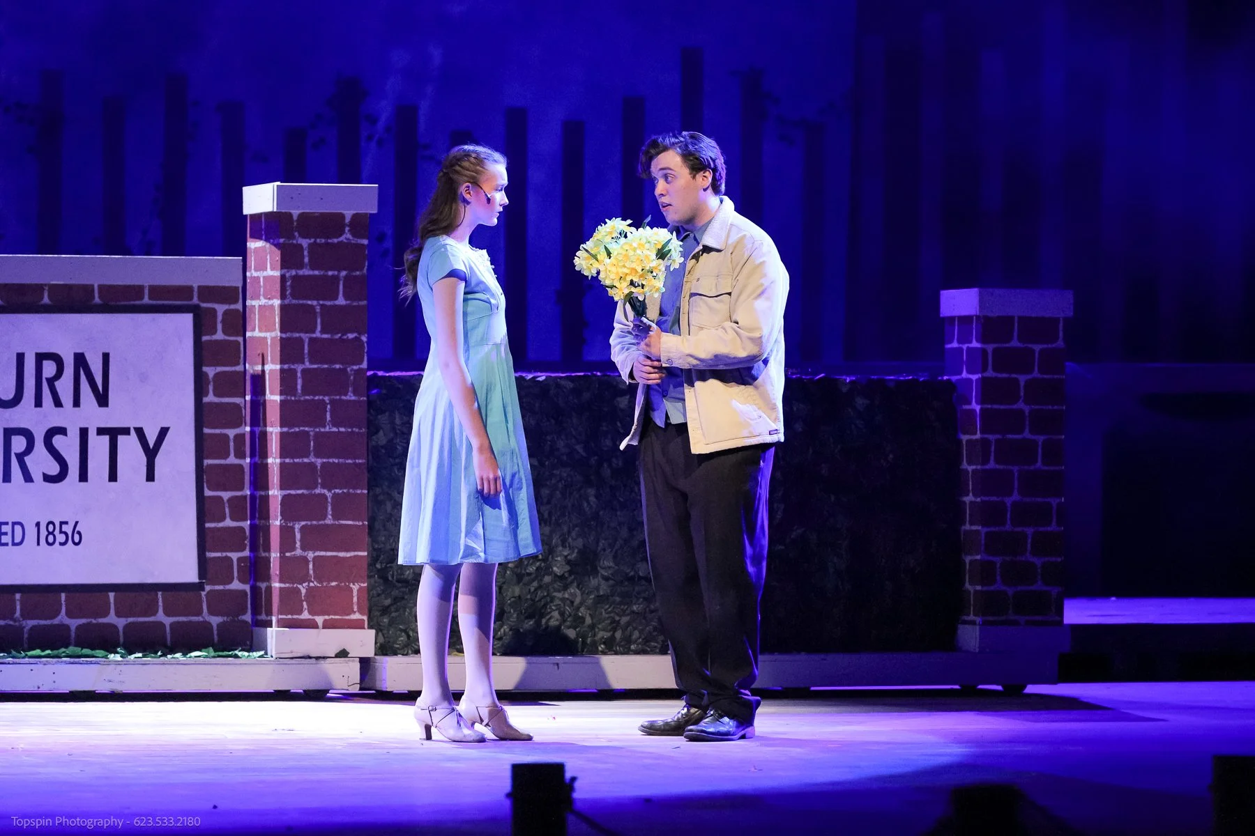 A young woman and a young man standing on stage in front of a sign reading "BURN UNIVERSITY". The woman is wearing a light-colored dress and heels, and the man is holding a bouquet of yellow flowers, wearing a light-colored jacket and dark pants. The